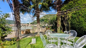 A garden patio with white metal chairs and a table, surrounded by greenery and a view of a town with a bridge and buildings. - Villa Rhôna (Andancette)
