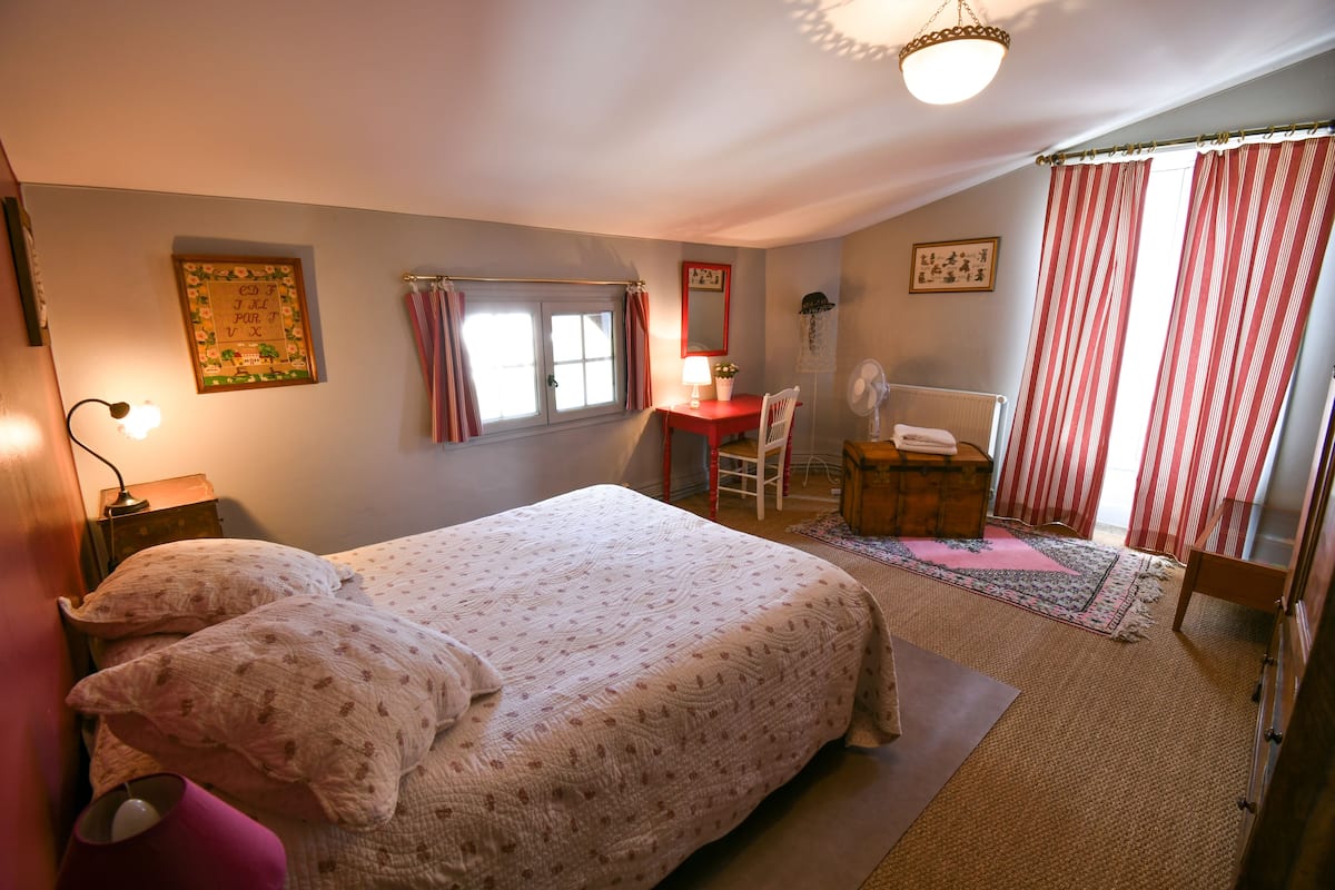 A bedroom with a bed, a desk, a chair, a fan, and a window with curtains.