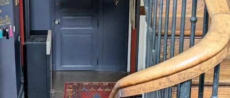 Una escalera de madera conduce a una puerta azul oscuro con un picaporte decorativo y un ventanal pequeño. Un tapete colorido se encuentra en el suelo frente a la puerta.