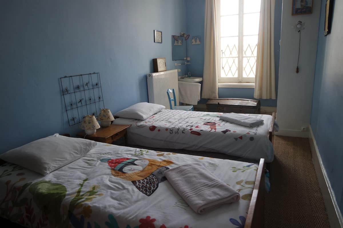 A bedroom with two beds, a radiator, a window with curtains, and a small table with a lamp.