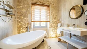 A modern bathroom with a freestanding bathtub, a stone wall, a window with blinds, a vanity with a mirror, and a towel rack.