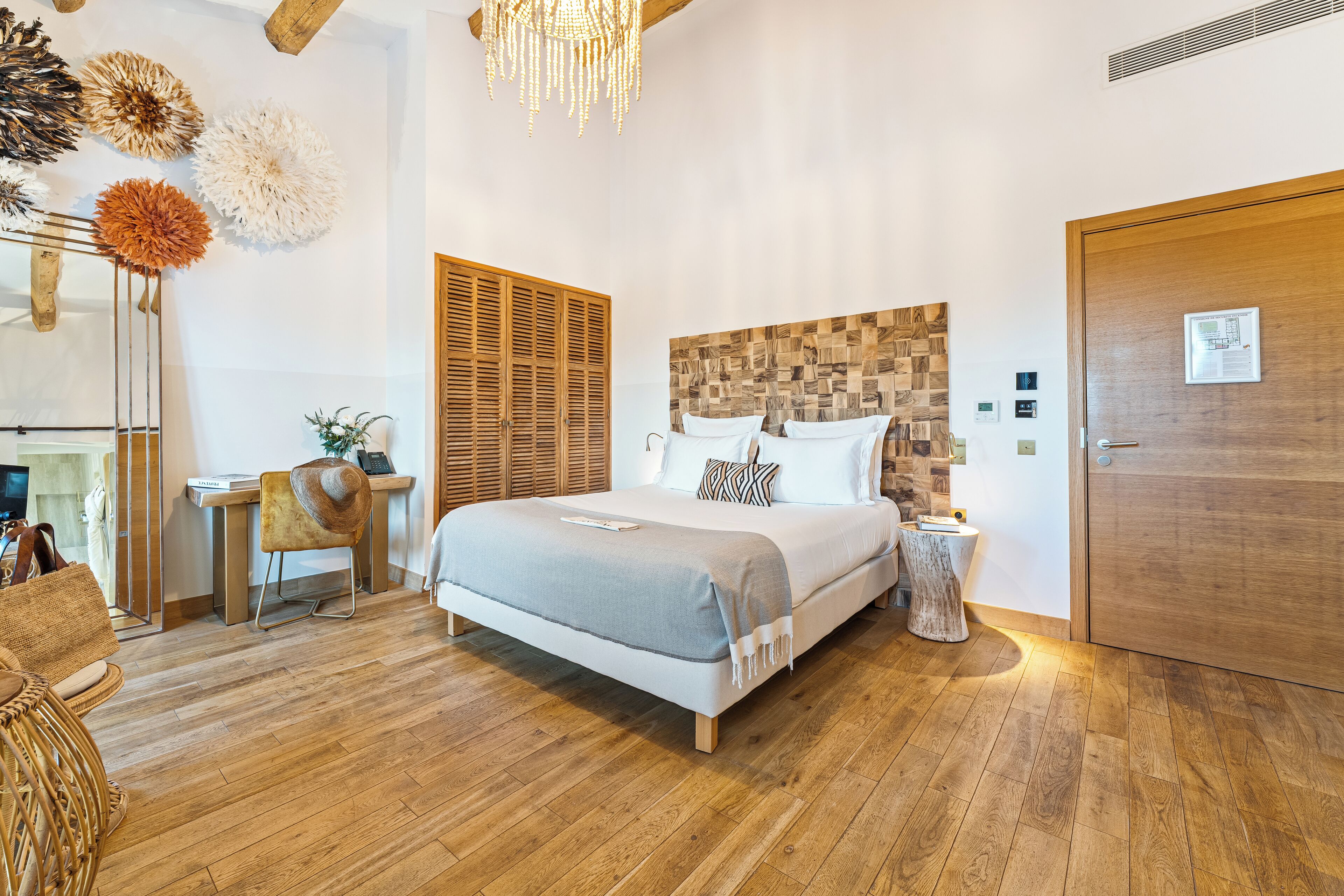 Un dormitorio con suelo de madera, una cama con manta gris, cabecero de madera, una mesita de noche y una puerta.