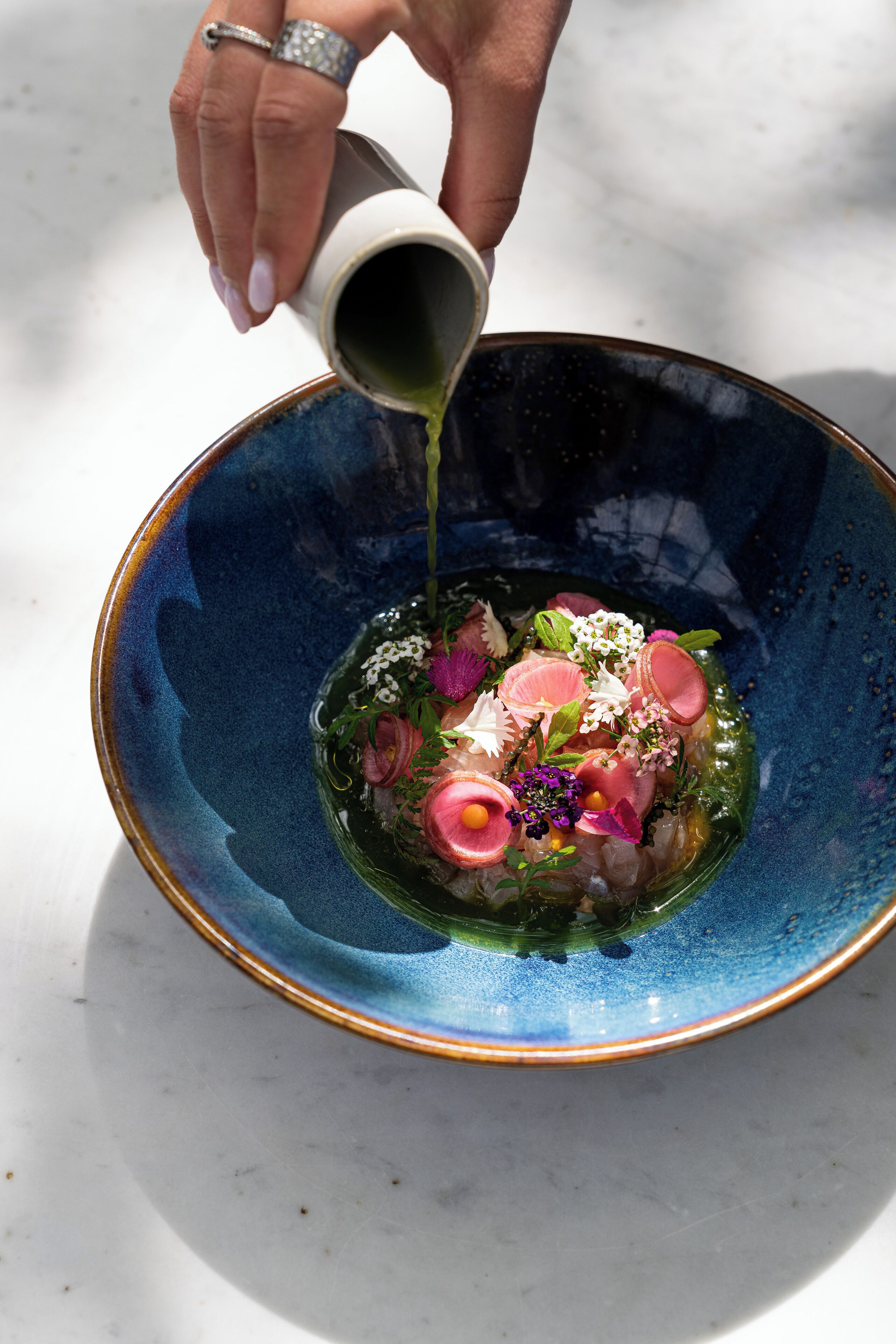 A hand pouring a liquid into a bowl containing food and garnishes.