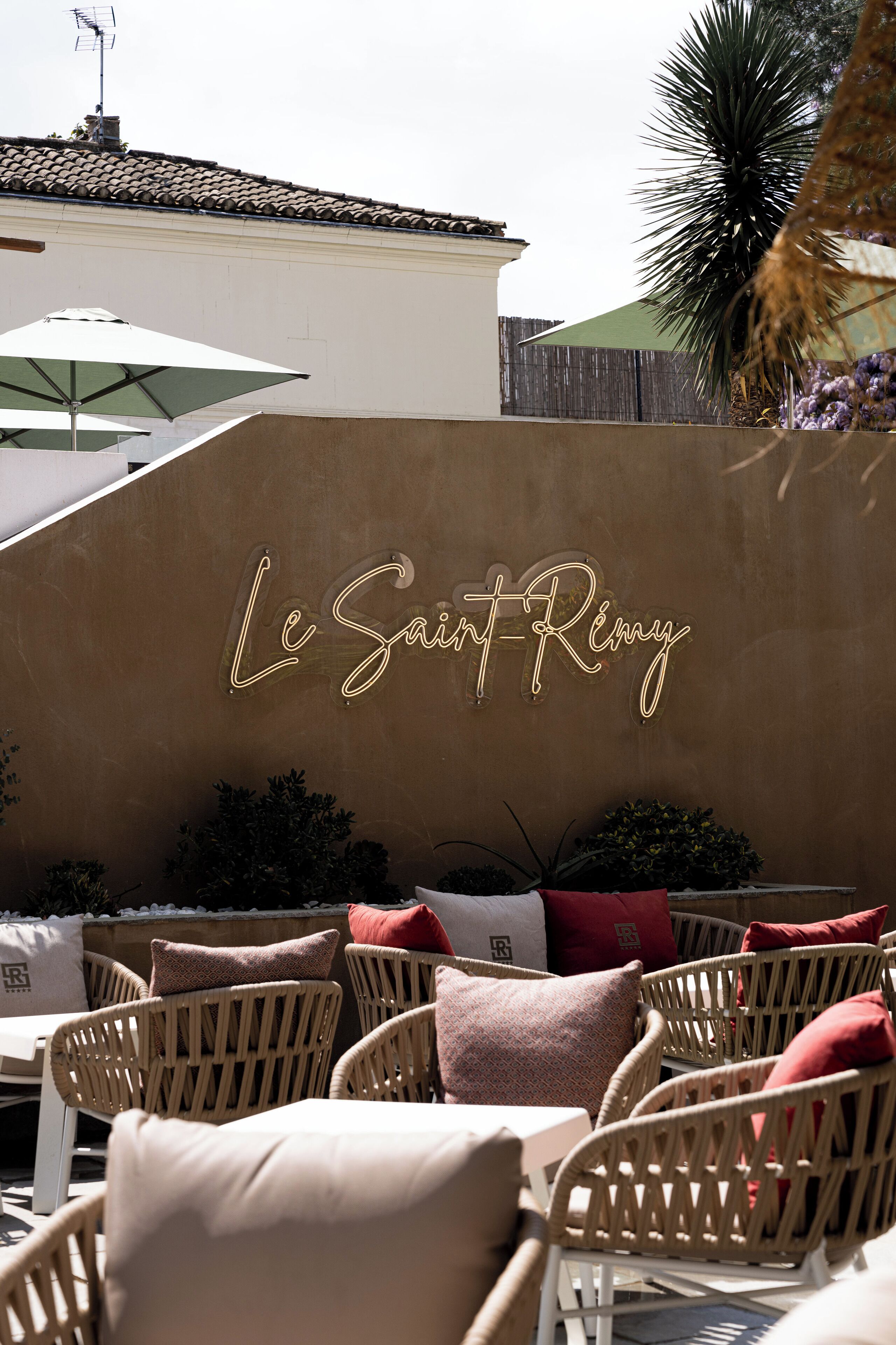 An outdoor seating area with wicker chairs and cushions, a neon sign reading "Le Saint-Rémy", and a tiled roof in the background.
