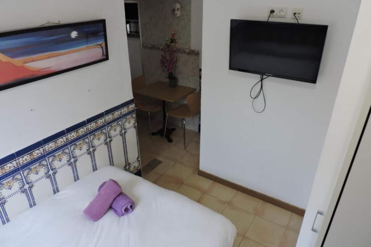 Habitación de hotel con una cama, un televisor montado en la pared, un cuadro y un pequeño comedor con mesa y sillas.
