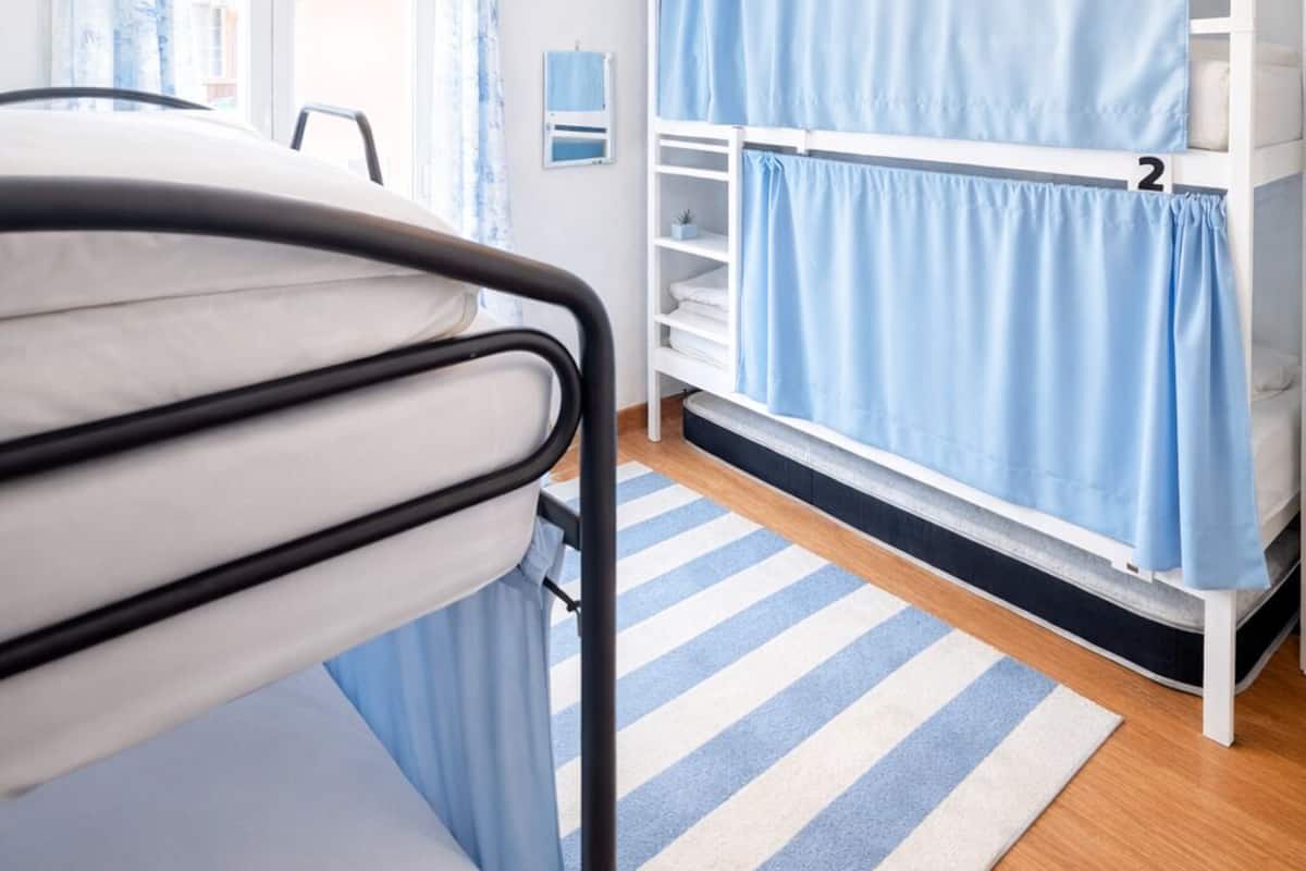 Una habitación con literas, un armario blanco, una cortina azul, un tapete a rayas y una lámpara colgante.