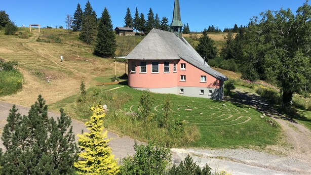 A church with a steeply pitched roof and a cross on top, surrounded by a grassy area with a circular path. - Berggasthaus Kandelhof (Simonswald)