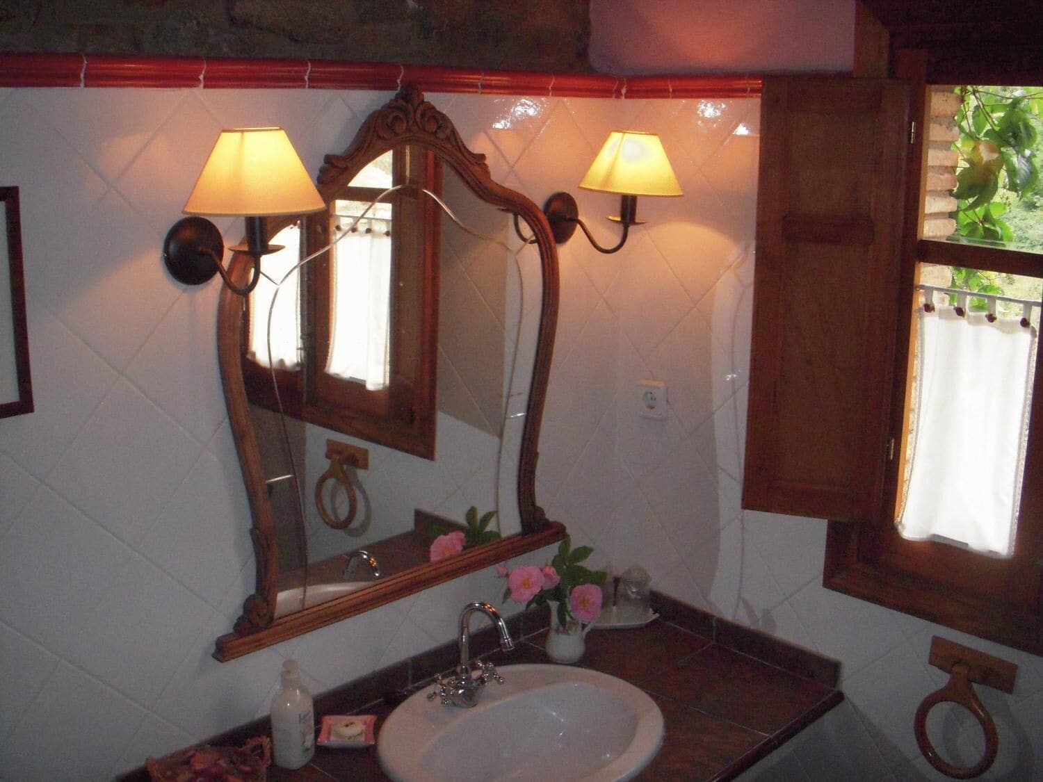Un baño con un espejo grande, dos lámparas de pared, un lavabo y una ventana con vistas a la vegetación.