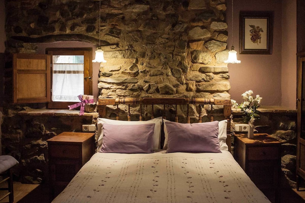 Een slaapkamer met een stenen muur, een houten hoofdbord, twee nachtkastjes, een bed met bloemenprint beddengoed, een raam met gordijn en een ingelijst schilderij aan de muur.