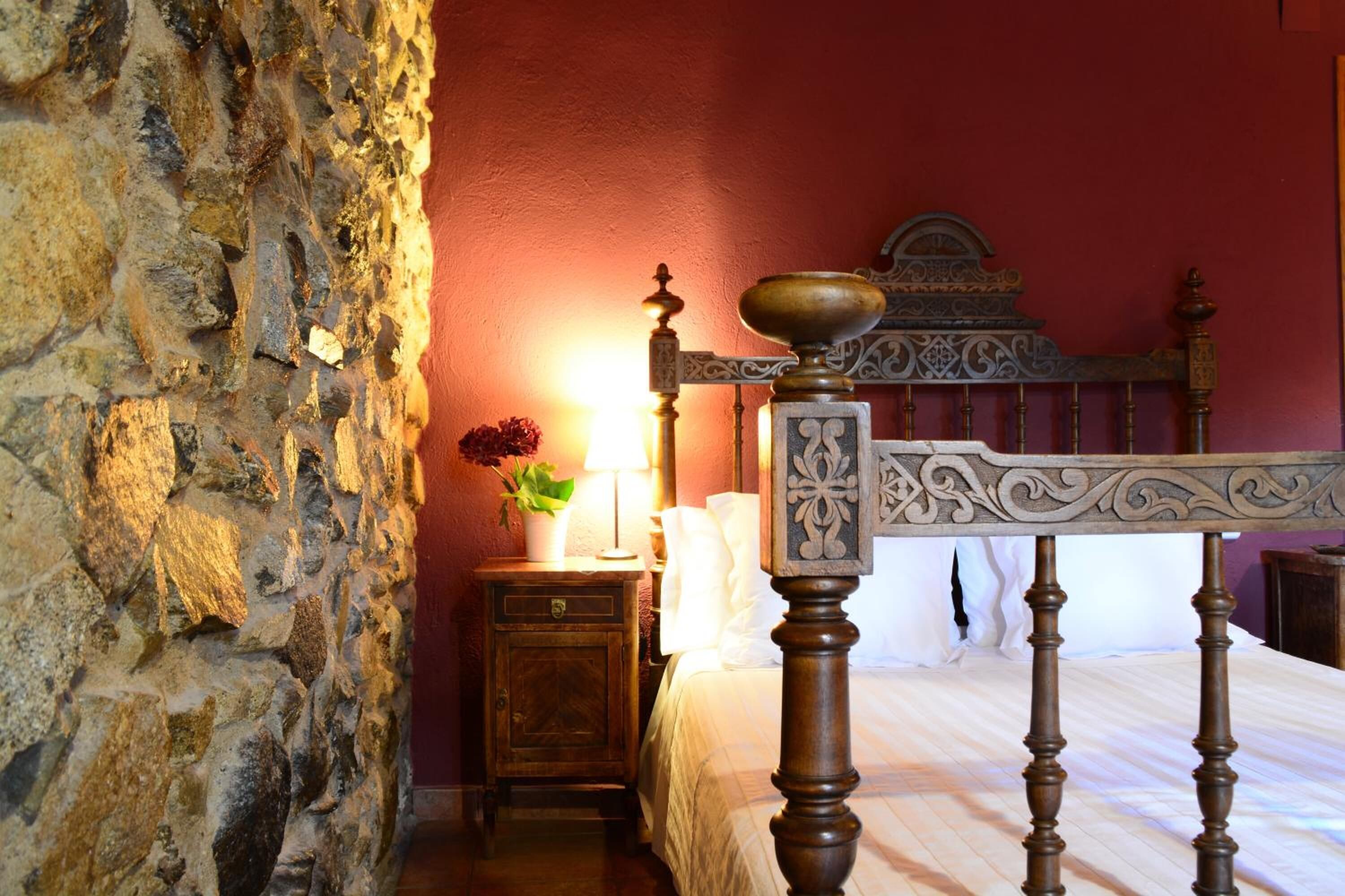 Un dormitorio con pared de piedra, cama de madera con cabecero decorado, mesita de noche con lámpara y planta en maceta.