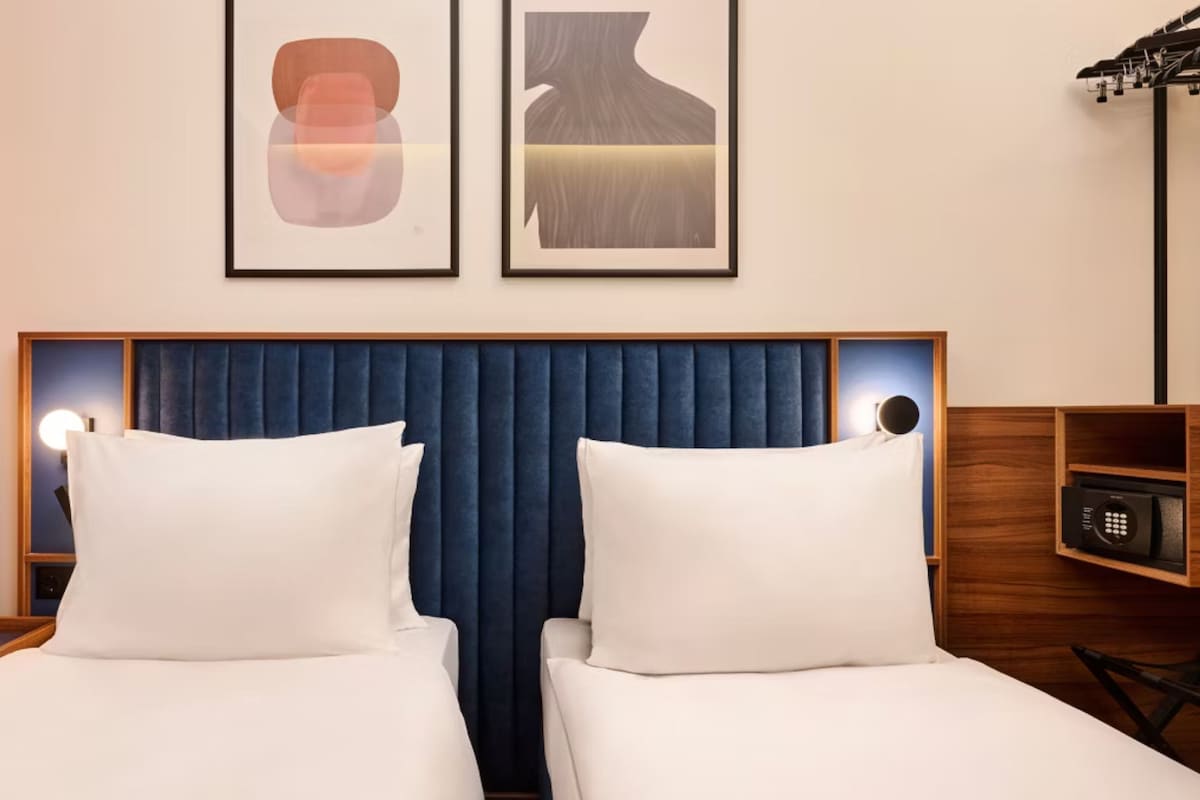 A hotel room with two beds, a blue headboard, a wooden side table with a remote control, and framed artwork on the wall.