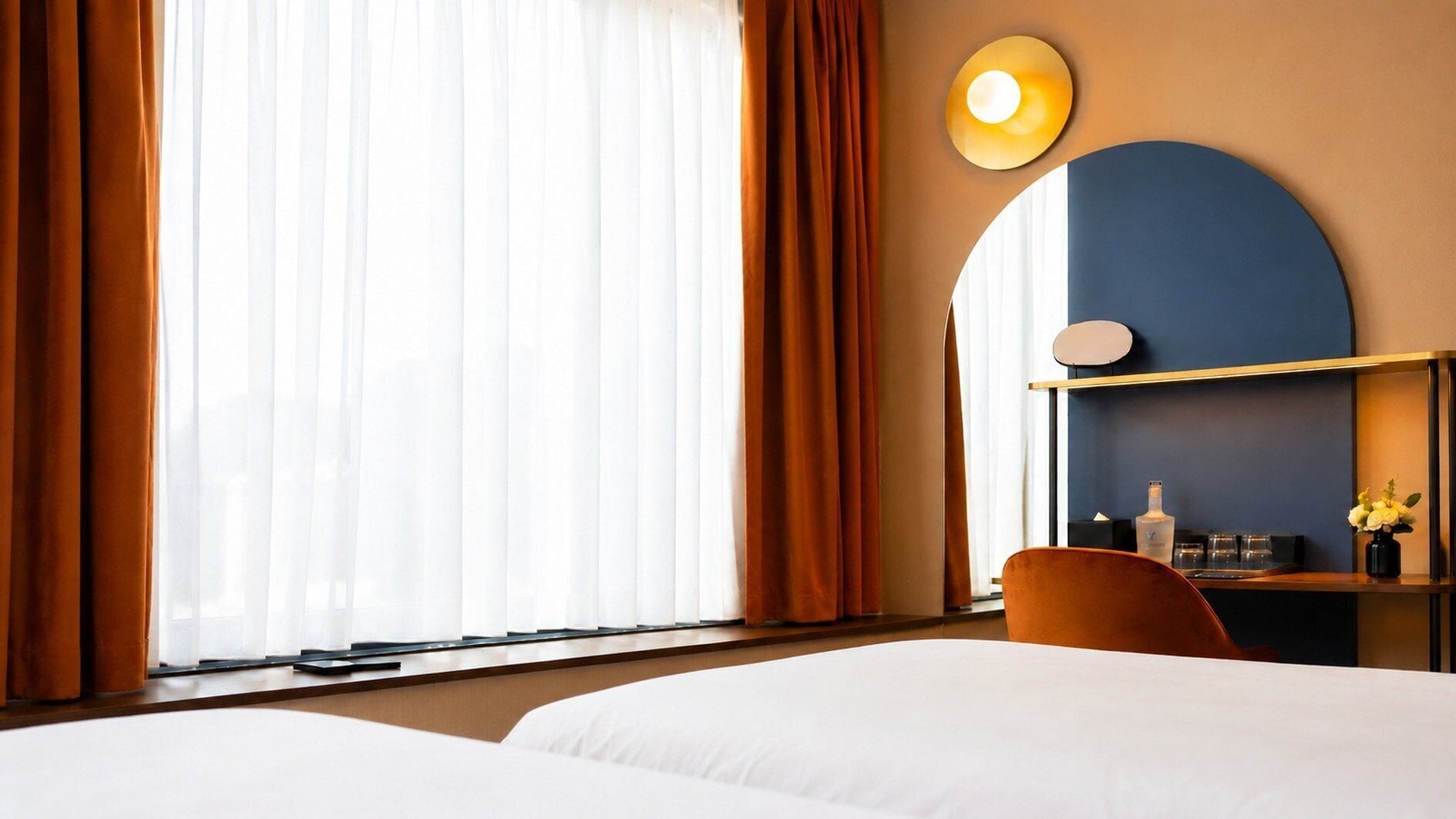 A hotel room with two beds, a desk with a chair, a round mirror, and a wall-mounted shelf with a bottle and glasses.
