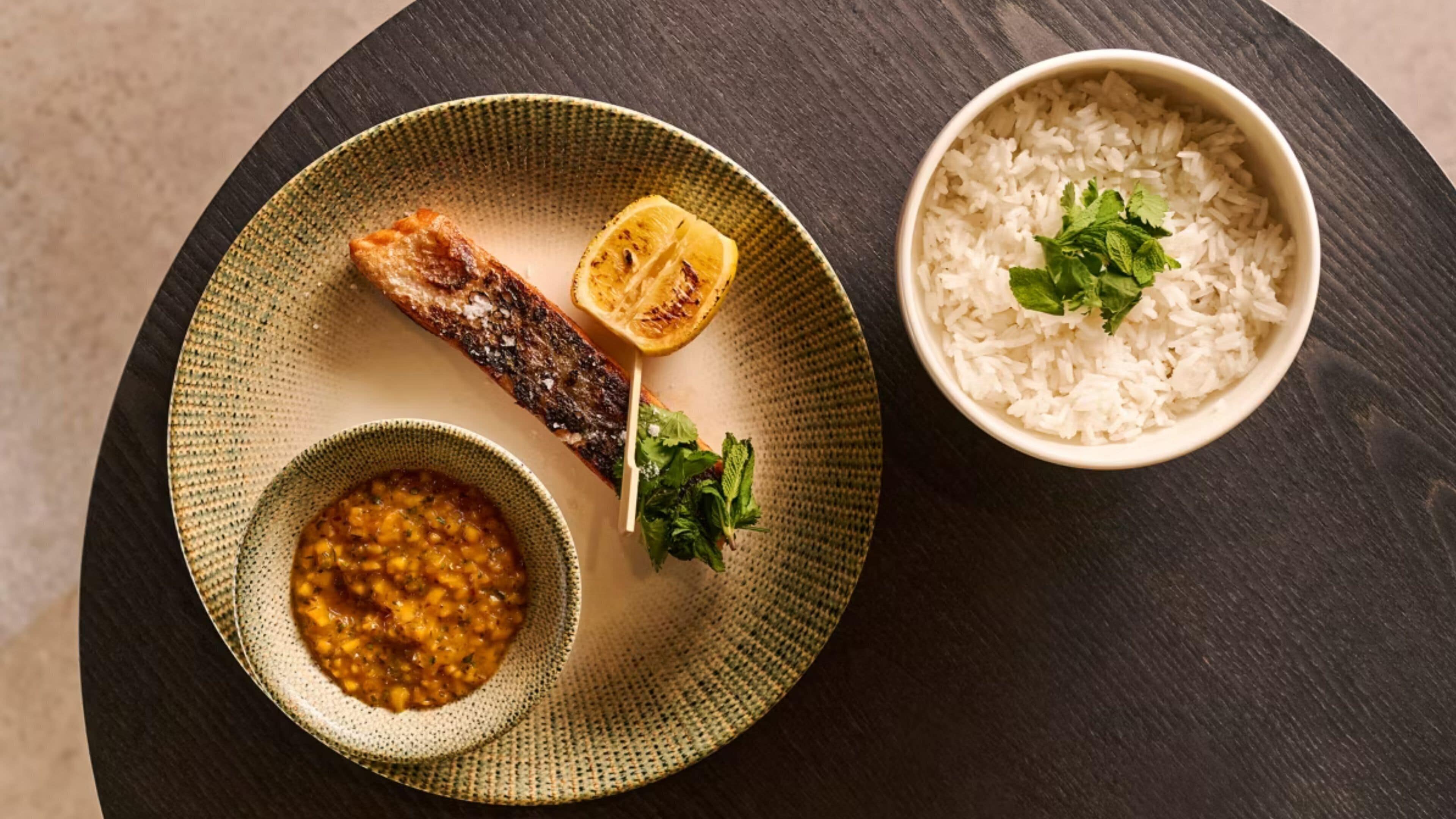 A plate with grilled meat, lemon, and a bowl of rice on a dark surface.