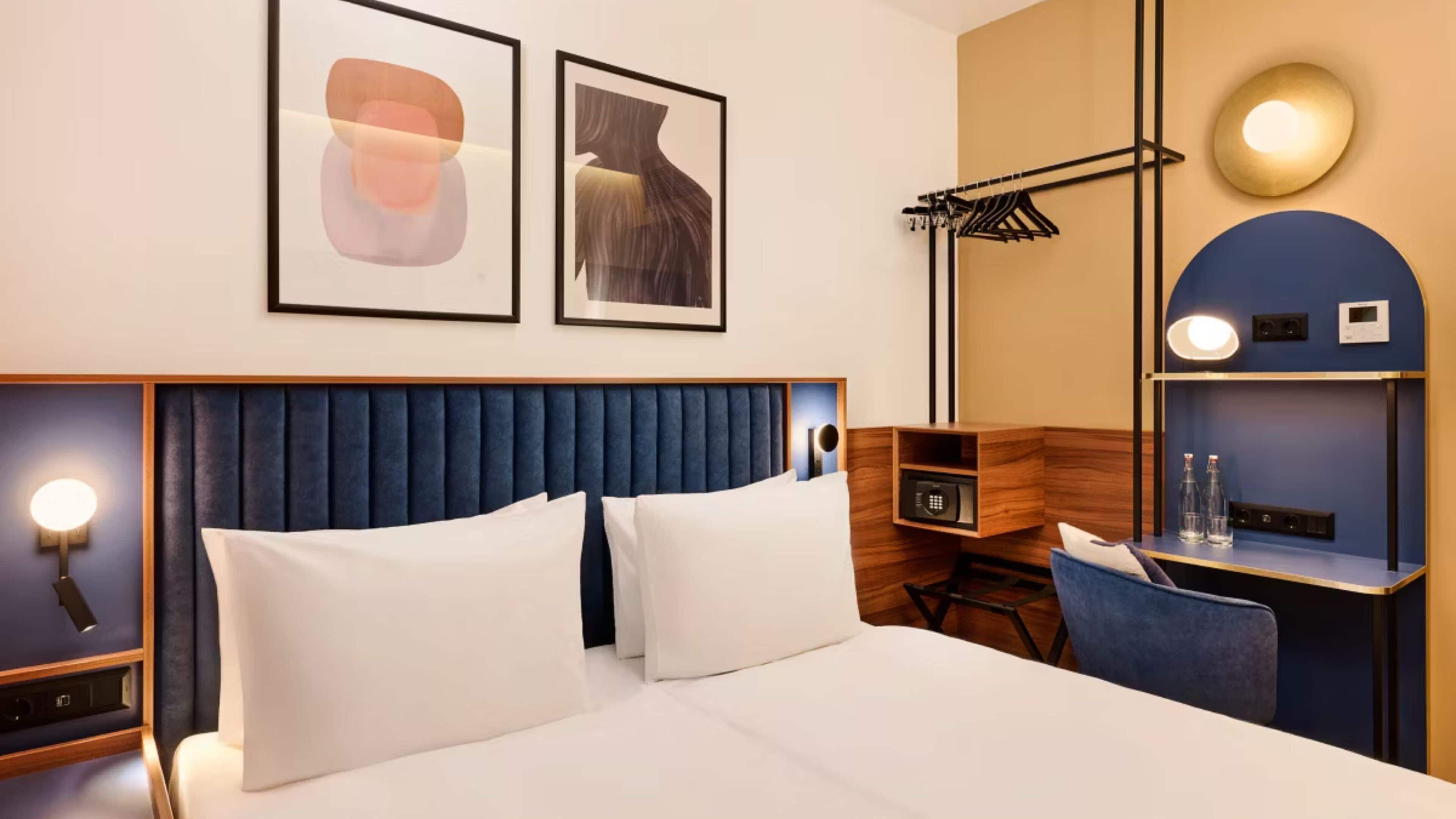 A hotel room with a large bed, a headboard, bedside tables, a desk, a chair, a wardrobe, and wall-mounted artwork.