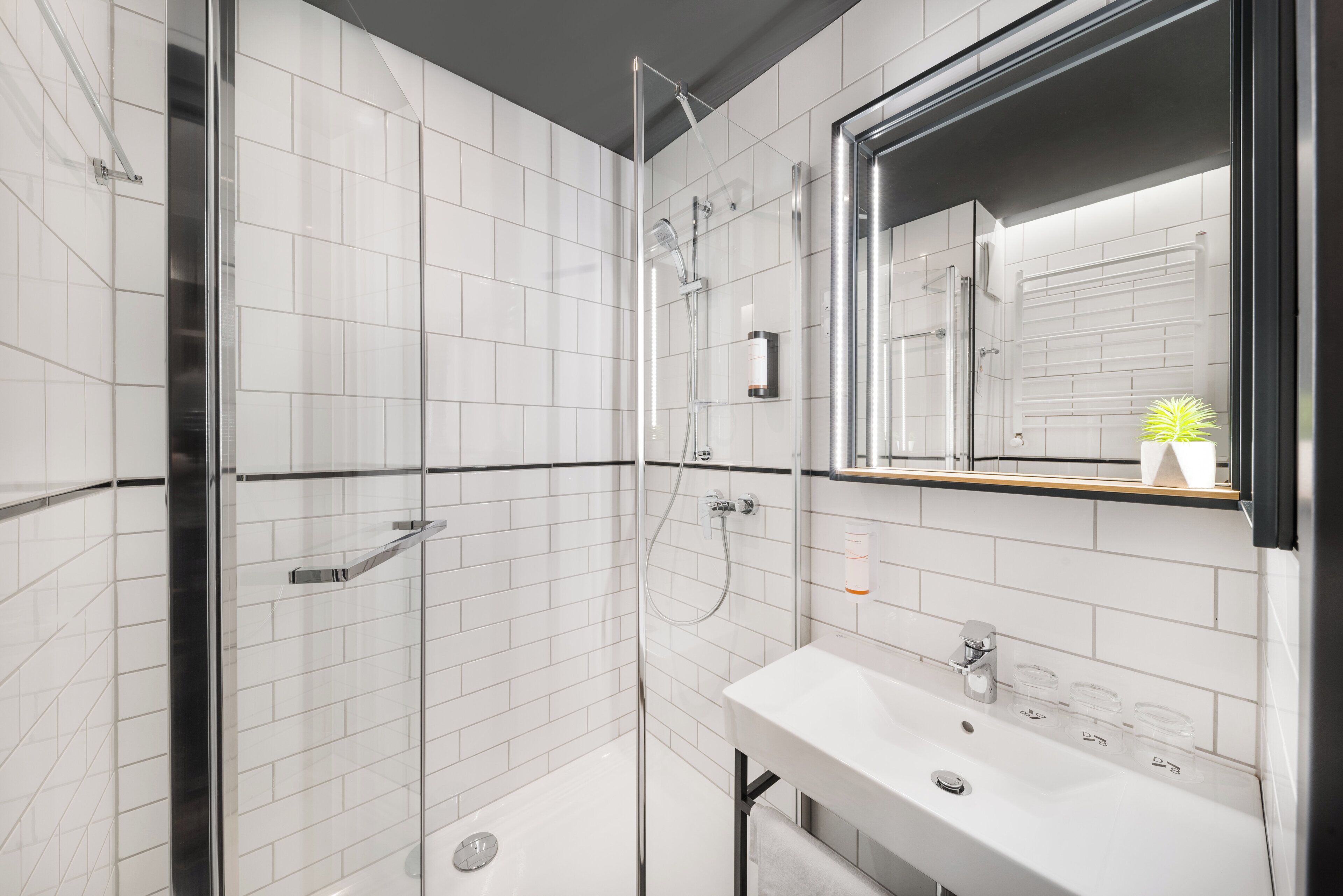 Bany modern amb rajoles blanques, un cubicle de dutxa de vidre, un lavabo amb mirall i un suport per a tovalloles.