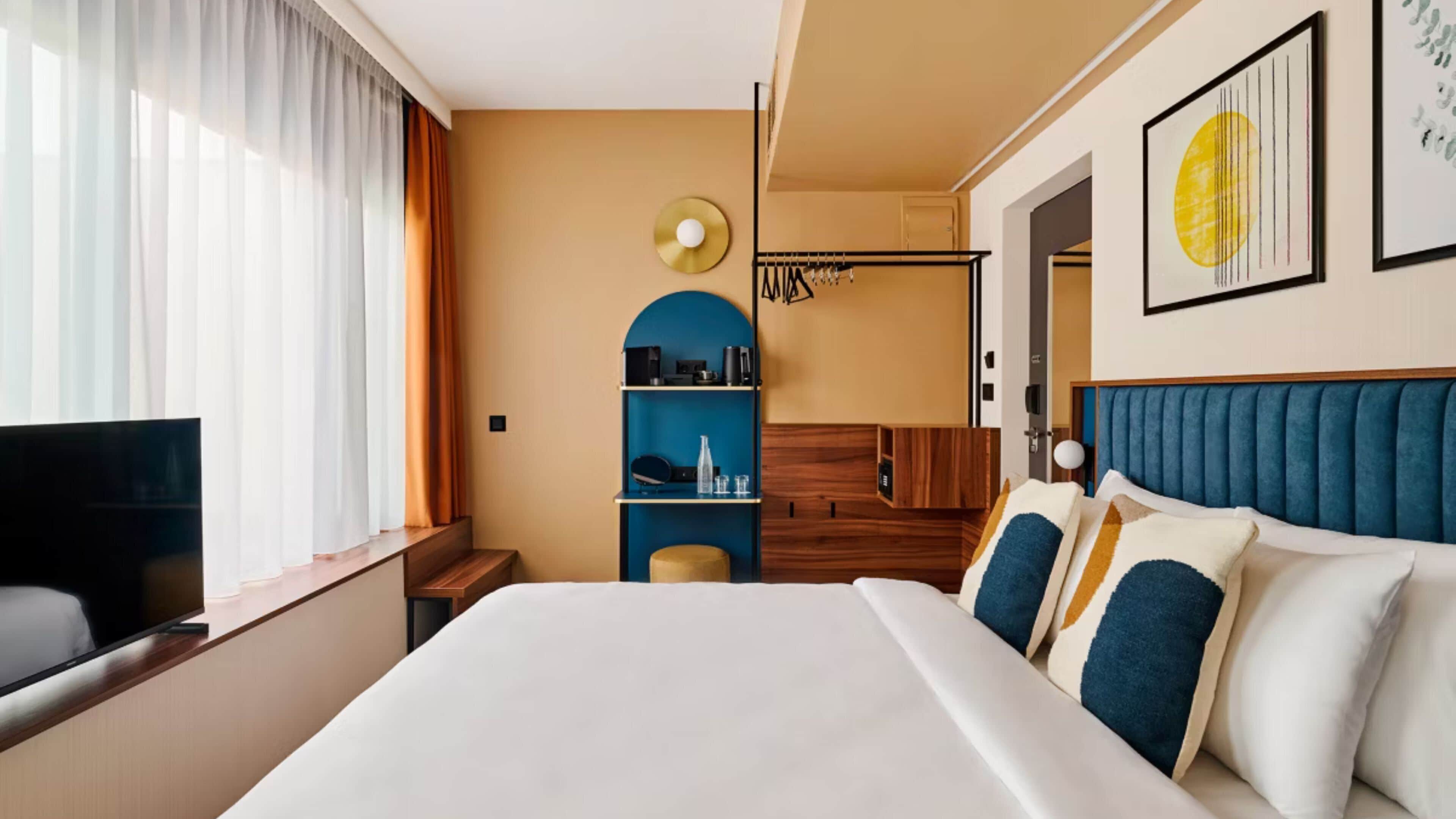 Een hotelkamer met een bed, een televisie, een kledingkast, een plank met spullen en een schilderij aan de muur.