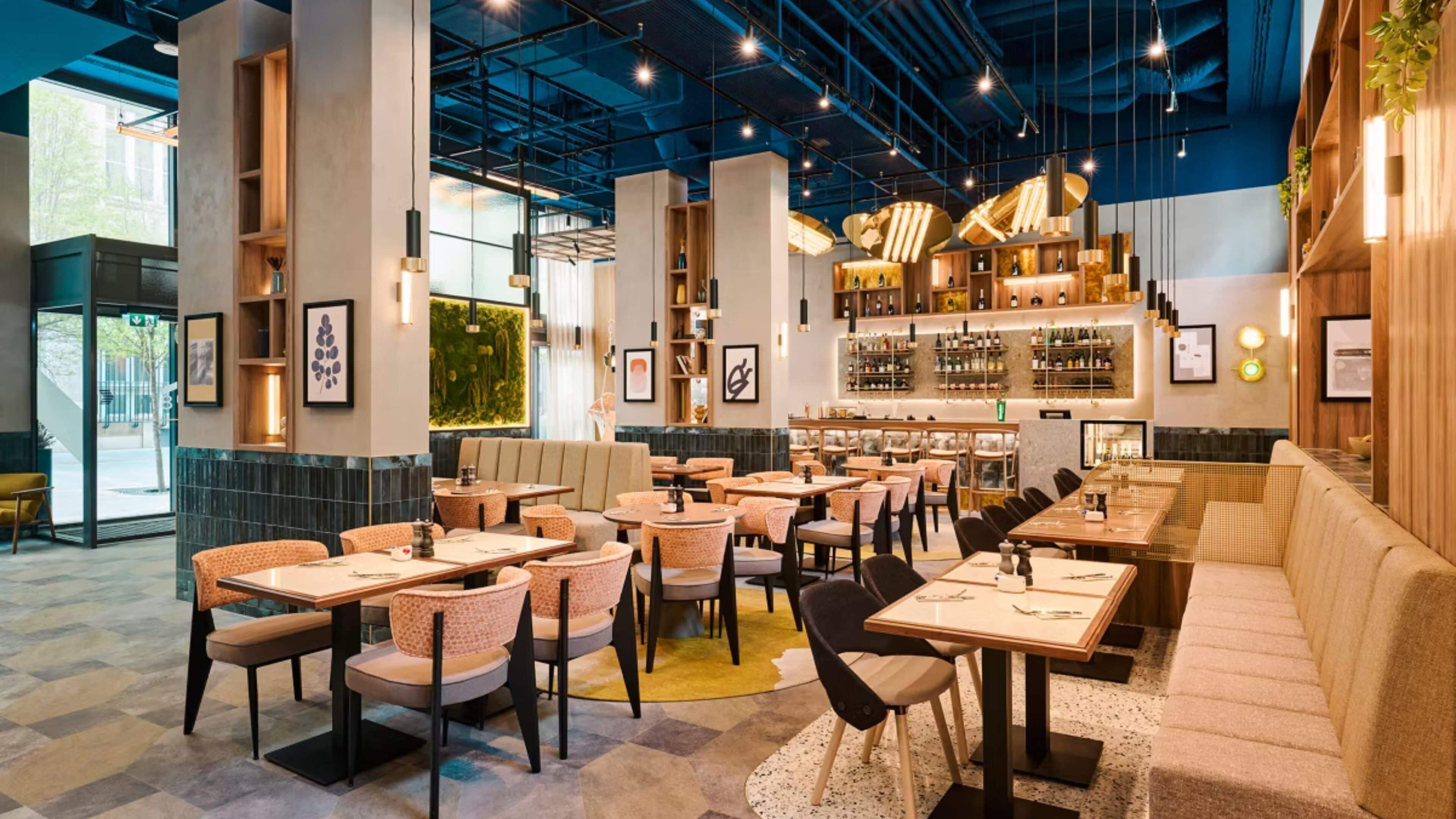 Un restaurante moderno con techos altos, vigas a la vista y una zona de bar con estantes llenos de botellas.