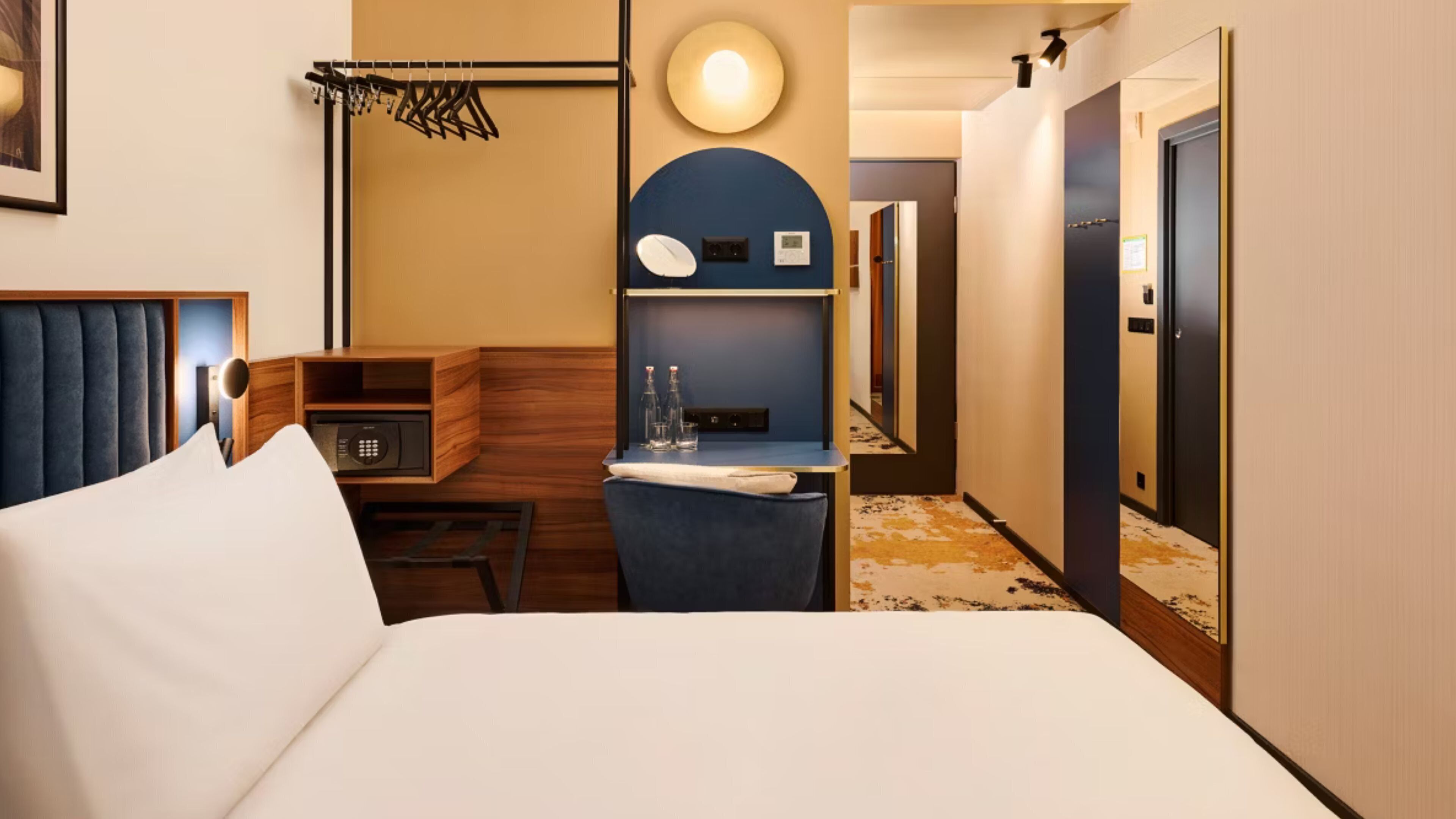 Habitación de hotel con cama, escritorio con silla, espejo, perchero y lámpara de pared.