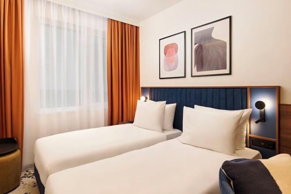 Habitación de hotel con dos camas, un ventanal con cortinas y dos cuadros en la pared.