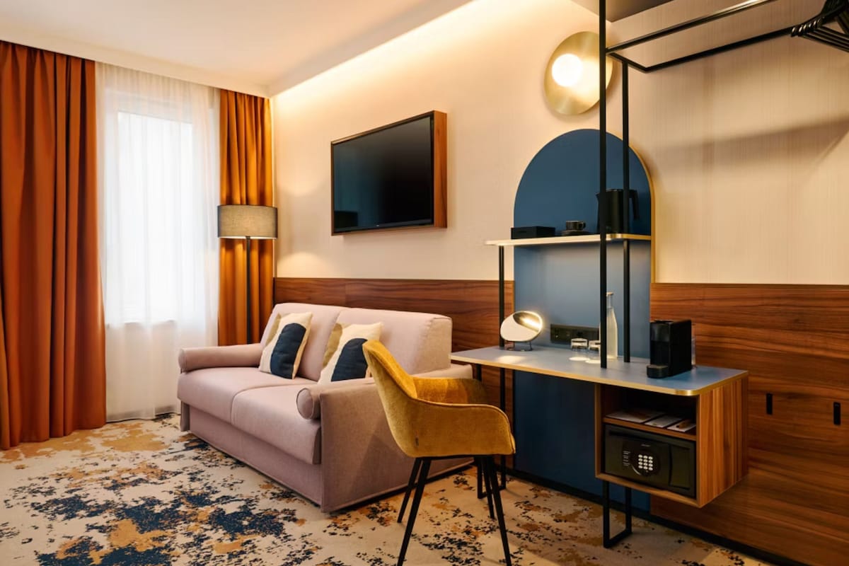 A hotel room with a sofa, a desk with a lamp, a chair, a TV, and a wall-mounted shelf.