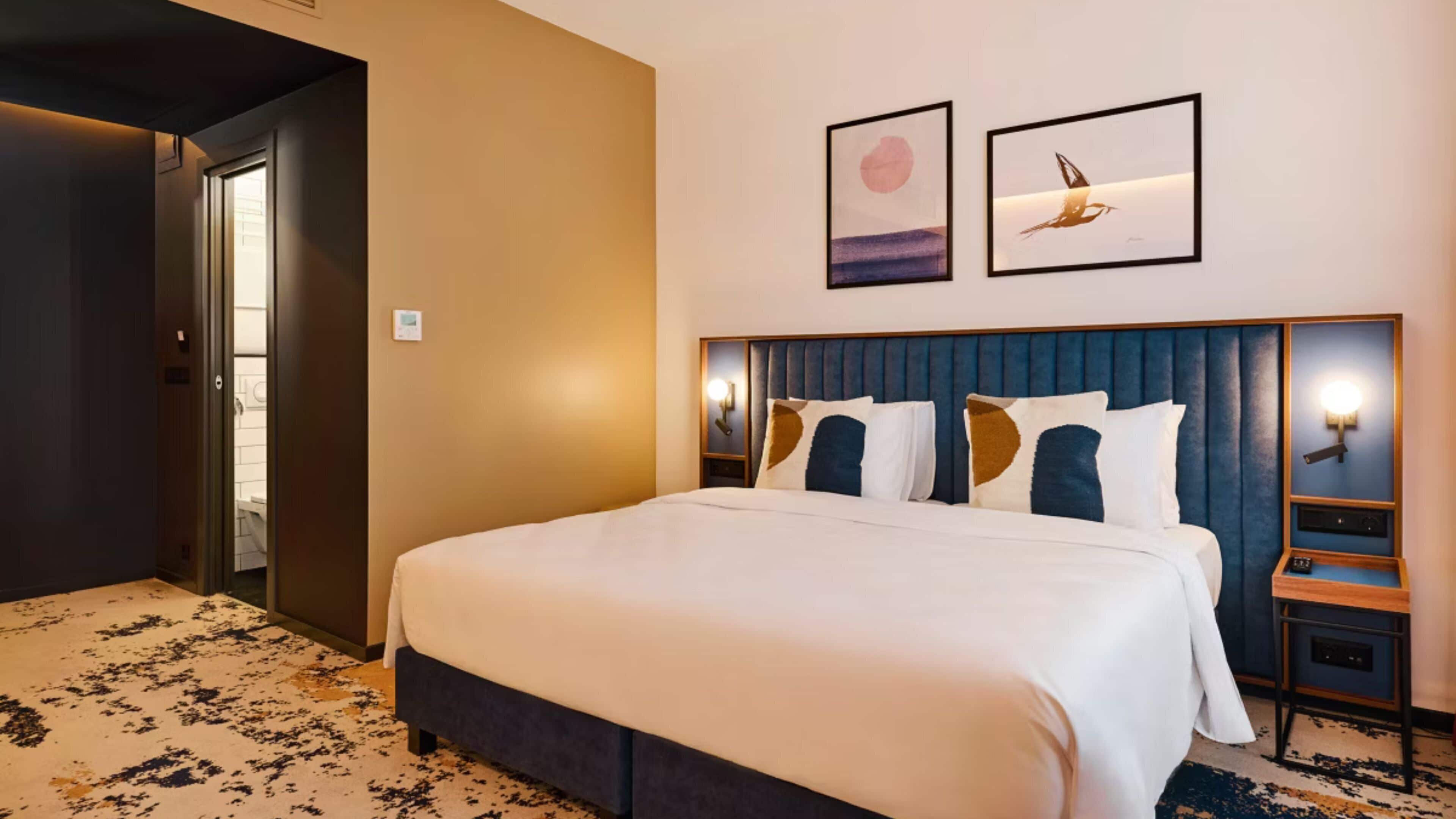 Quarto de hotel com uma cama grande, cabeceira com iluminação embutida, duas obras de arte na parede e piso coberto por carpete.