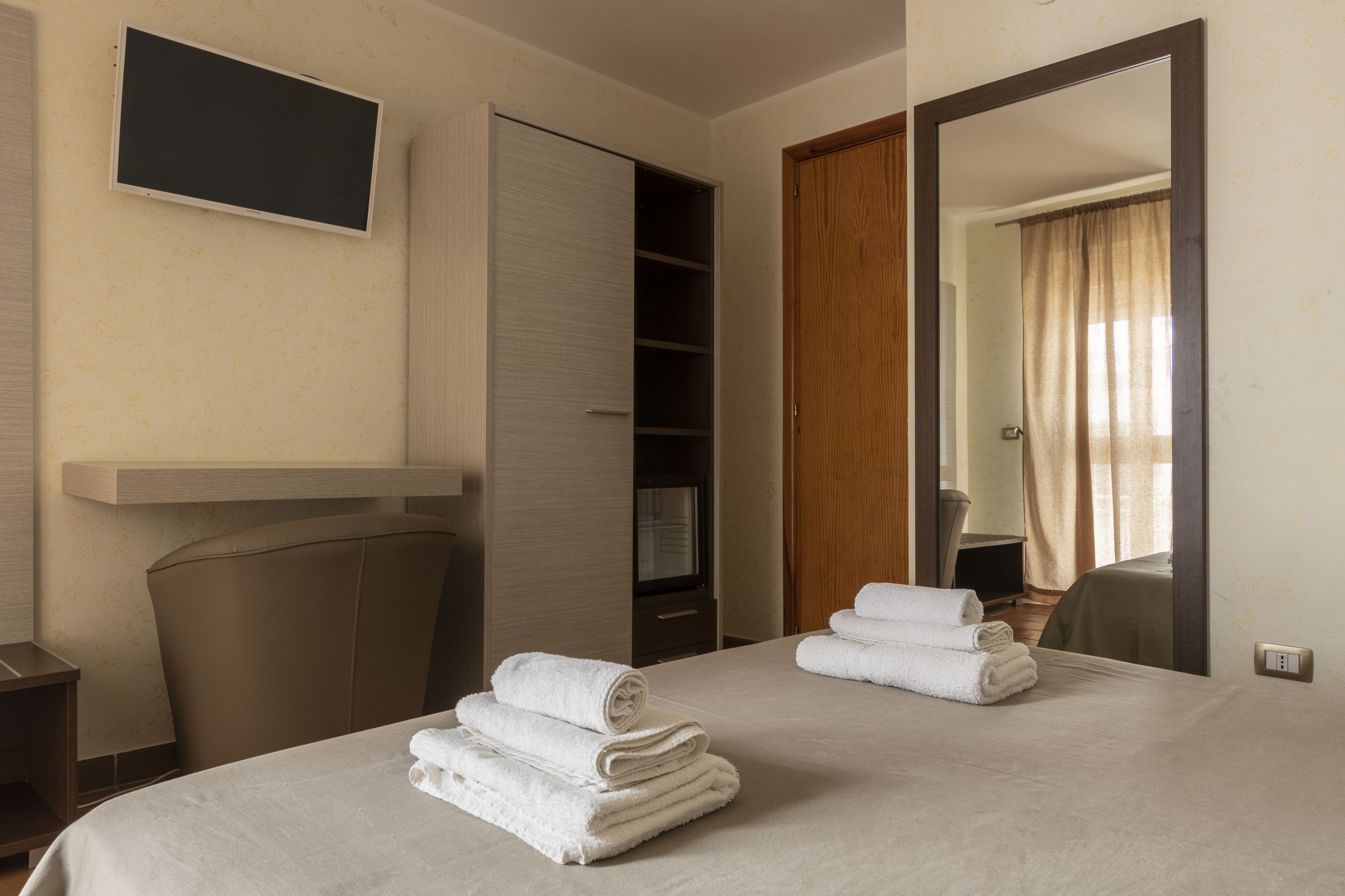 A hotel room with a bed, two rolled towels, a TV, a wardrobe, and a mirror reflecting a bathroom.