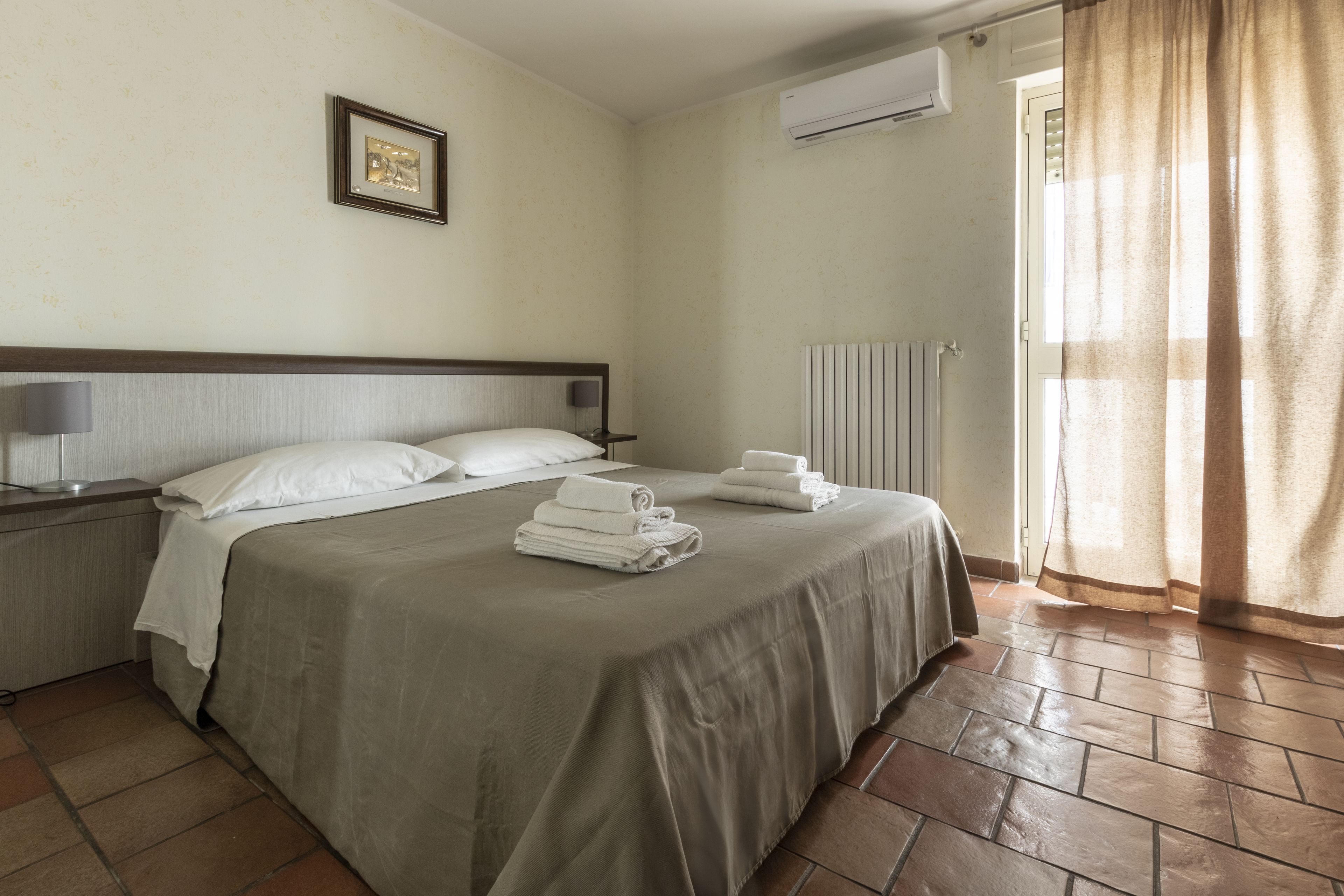 Habitación de hotel con cama, mesita de noche, lámpara, un cuadro en la pared y ventana con cortinas.
