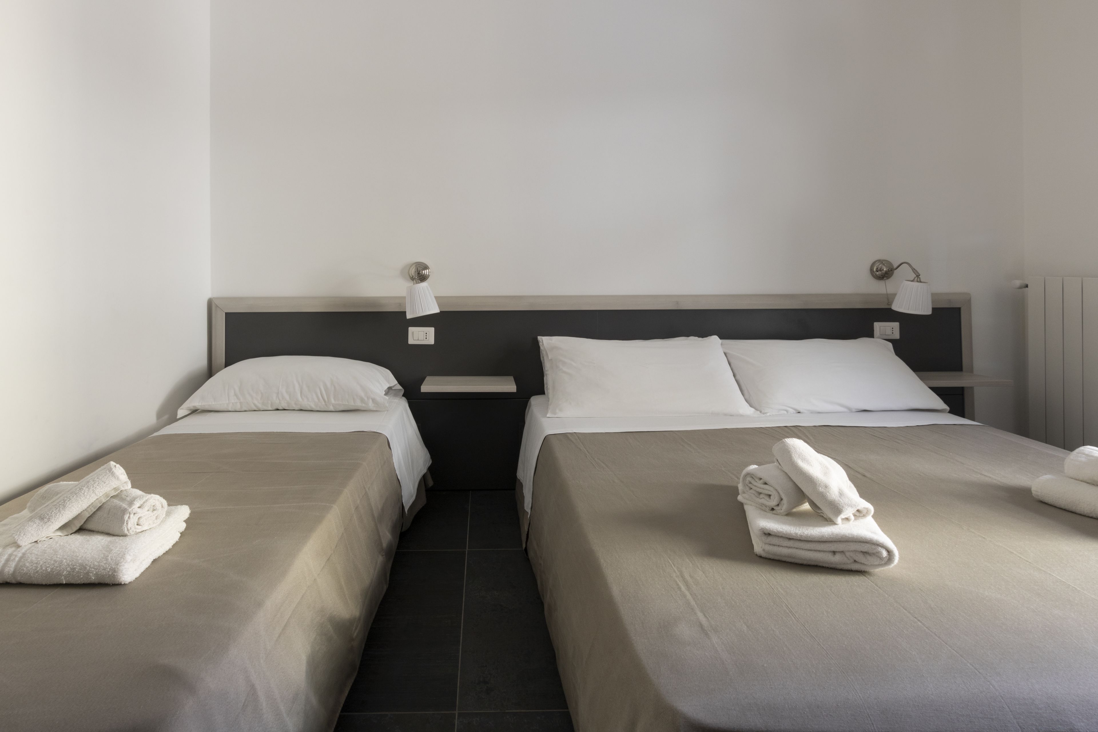 A hotel room with two single beds, each with a pillow and a folded towel. There are bedside tables with lamps and a wall-mounted light fixture.