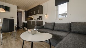 Una sala moderna con un sofá gris, una mesa de centro blanca y una silla negra. Se observa un área de cocina con armarios de madera oscura y un horno empotrado.