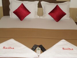 Una cama bien hecha con dos almohadas rojas, una manta marrón y dos toallas con el nombre "Saiba" bordado.