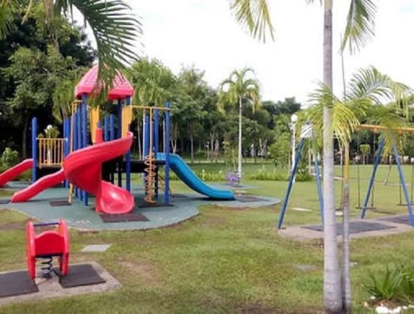 A playground with slides and swings, surrounded by grass and palm trees. - KK City HomeSuite at KK City Center 2 (Kota Kinabalu)