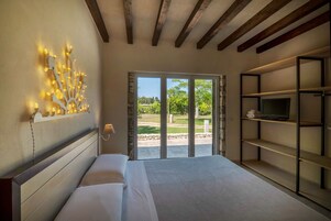 A bedroom with a large bed, a wall-mounted light fixture, a television on a shelf, and a view of a garden through a glass door.