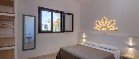 A bedroom with a bed, two bedside tables with lamps, a large window, a wall-mounted mirror, and a wall light fixture.