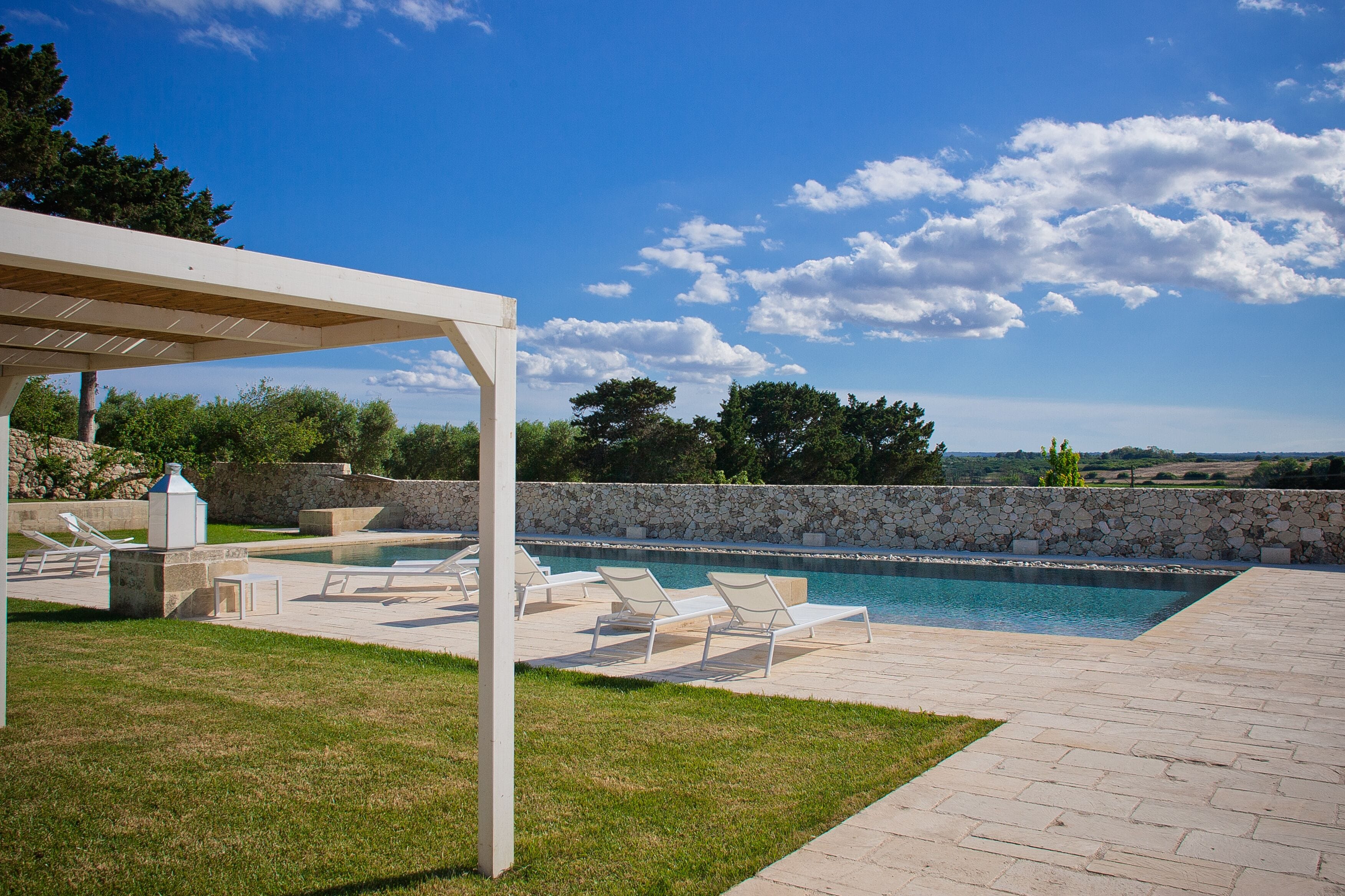 A swimming pool area with lounge chairs, a pergola, and a stone wall.