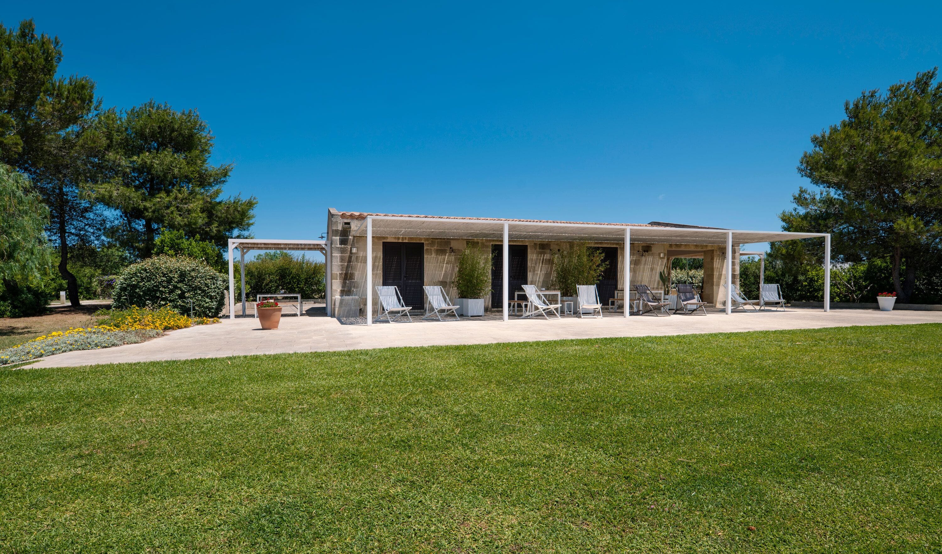 A covered outdoor seating area with multiple lounge chairs and tables, surrounded by a well-maintained lawn and trees.