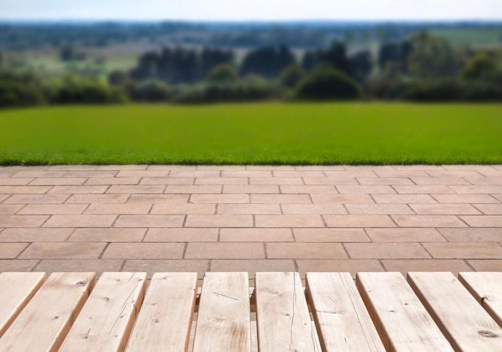 A paved patio with wooden tables and a view of a green field and trees in the distance.