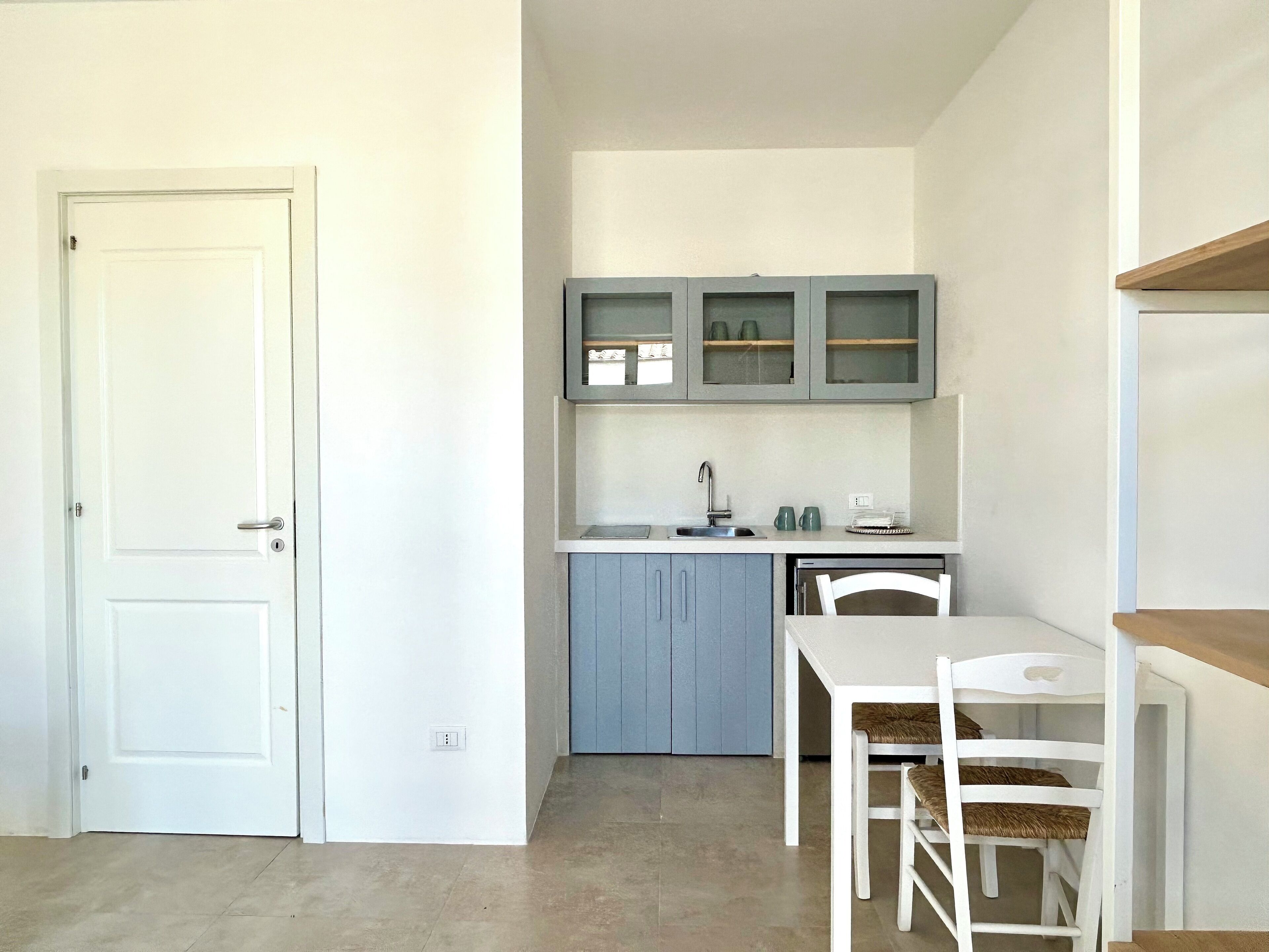 A compact kitchen with a white door, a sink, a small dining table with chairs, and a cabinet with glass doors.