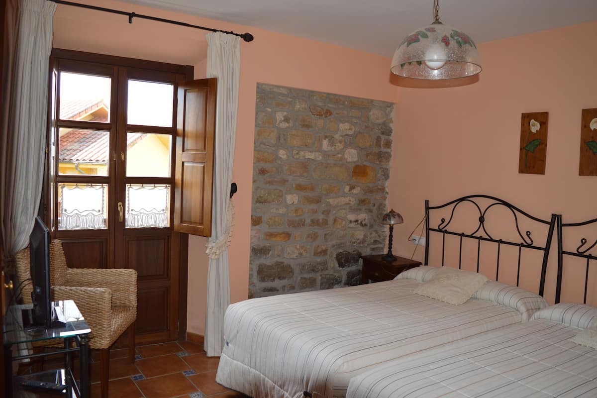 Ein Schlafzimmer mit Bett, Steinwand, Fenster mit Vorhängen, einem Korbstuhl und einer Lampe.