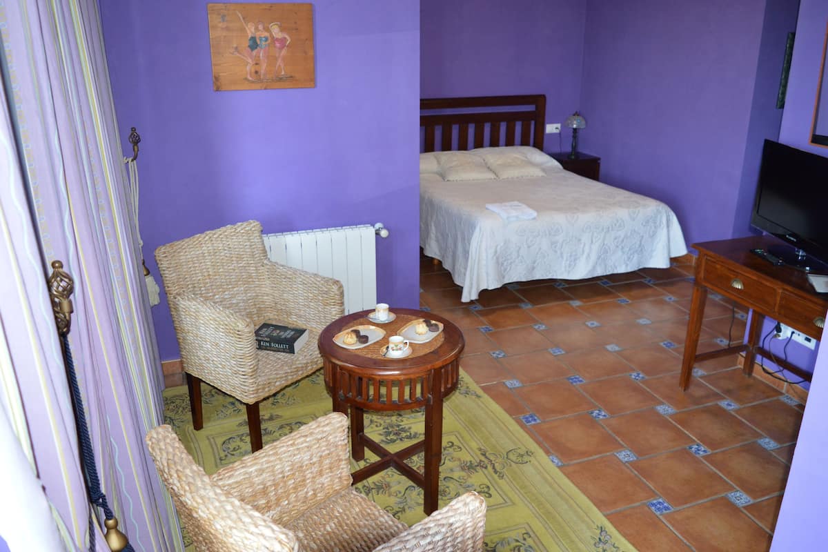 Ein Zimmer mit einem Bett, einem Sessel, einem kleinen Tisch mit Teetassen, einem Heizkörper und einem Fernseher.
