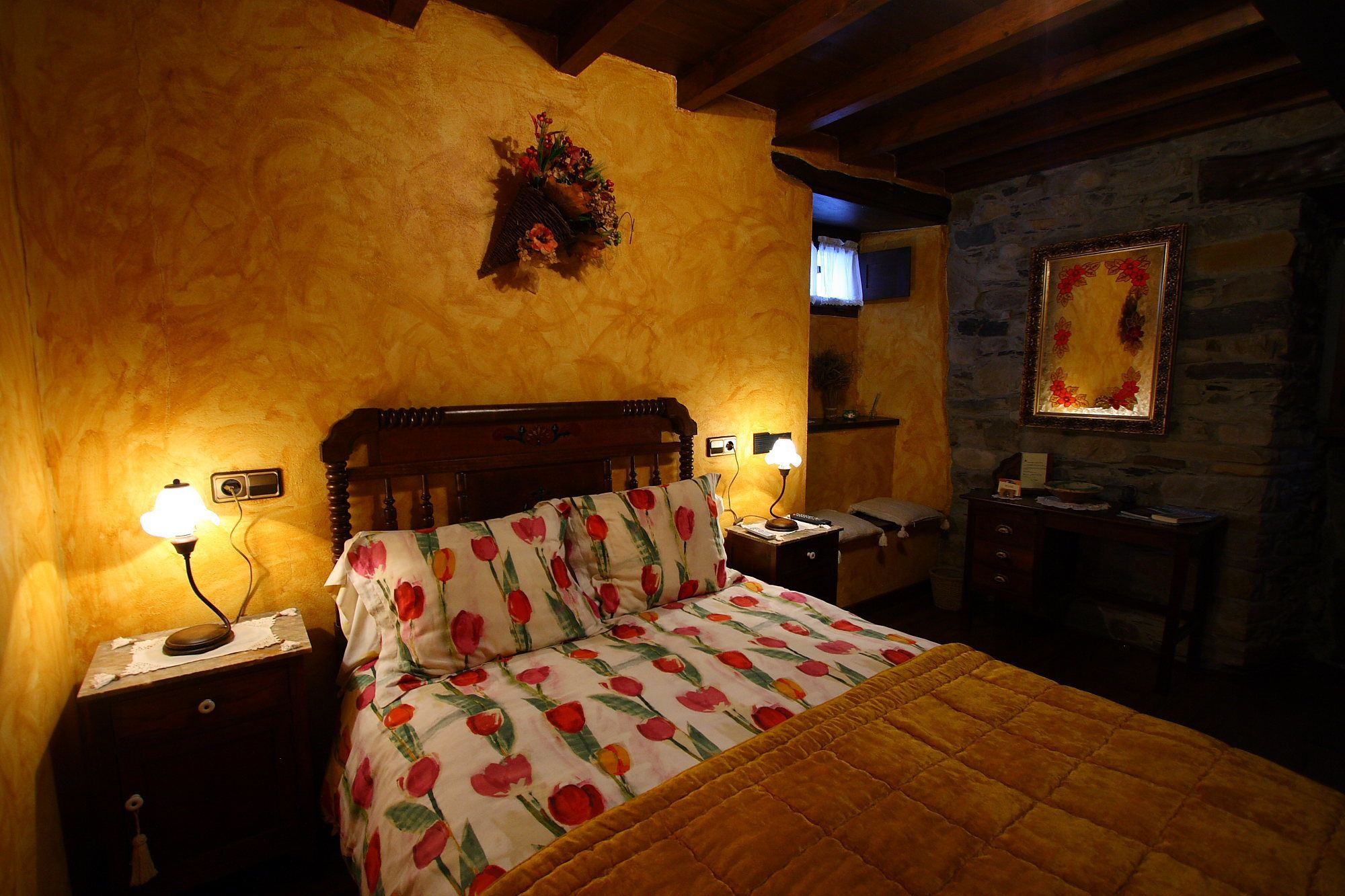 Ein gemütliches Schlafzimmer mit einem hölzernen Bett, floralem Bettbezug, einem Nachttisch mit Lampe, einer Kommode und einer Steinwand mit Spiegel.