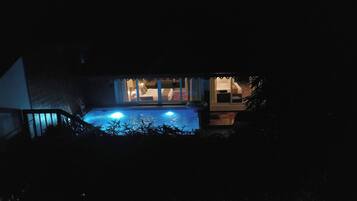 Vista noturna de uma área de piscina com deck coberto e um edifício ao fundo.