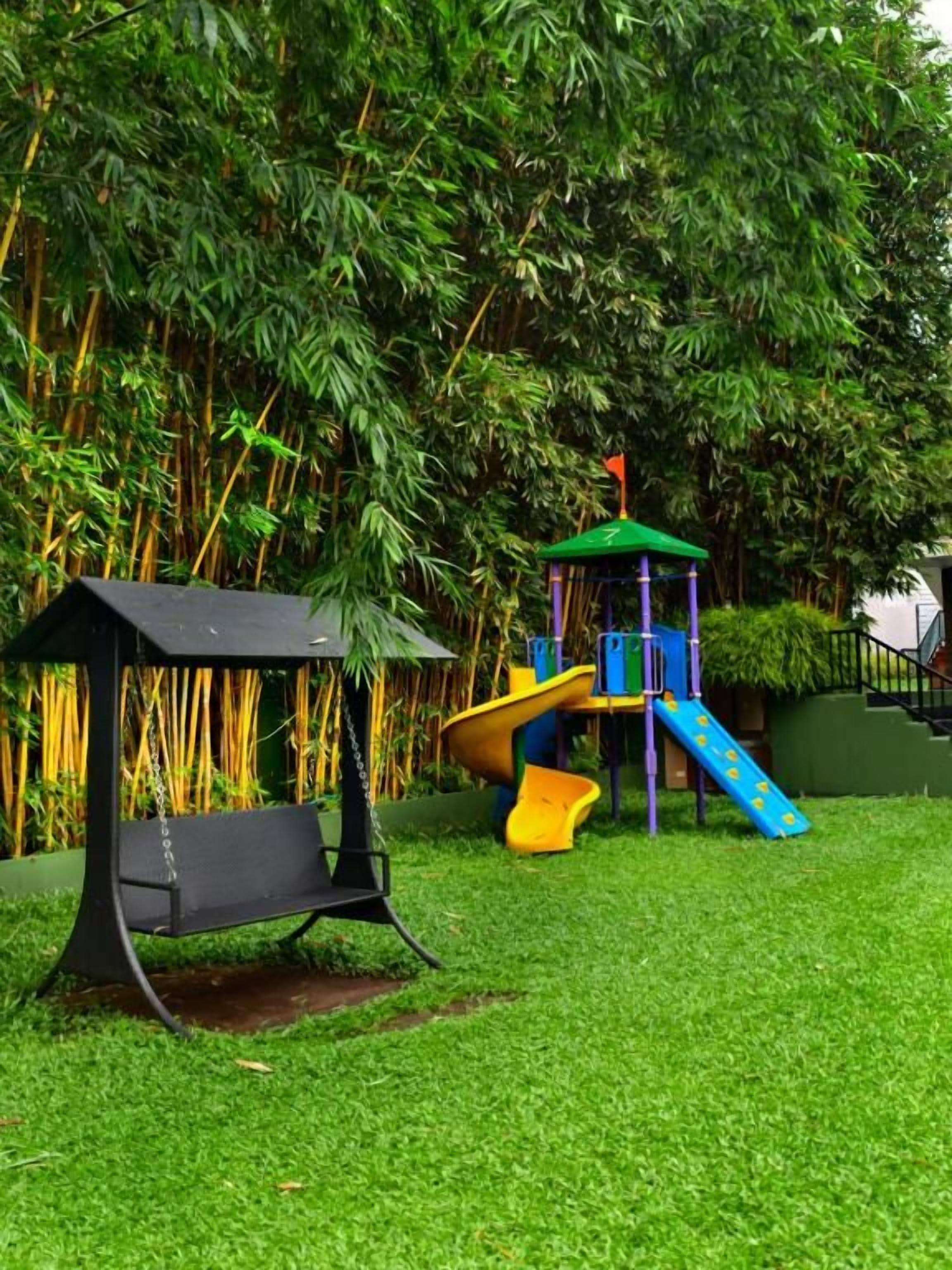A playground with a slide and a swing set, surrounded by lush greenery.