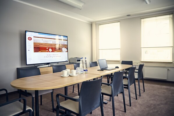 Une salle de conférence avec une longue table, des chaises, un ordinateur portable, un écran de projection et une fenêtre avec des stores.