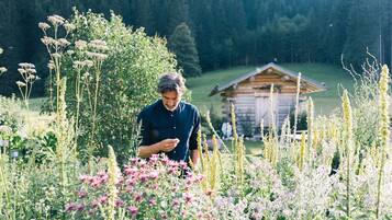 A person in a field with various plants and a wooden cabin in the background.