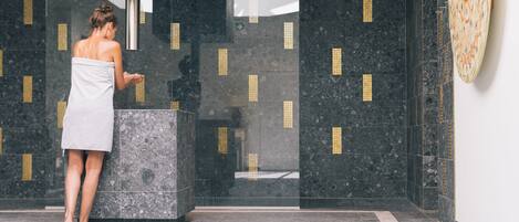 A woman in a towel standing in a modern bathroom with dark marble walls and a large mirror.