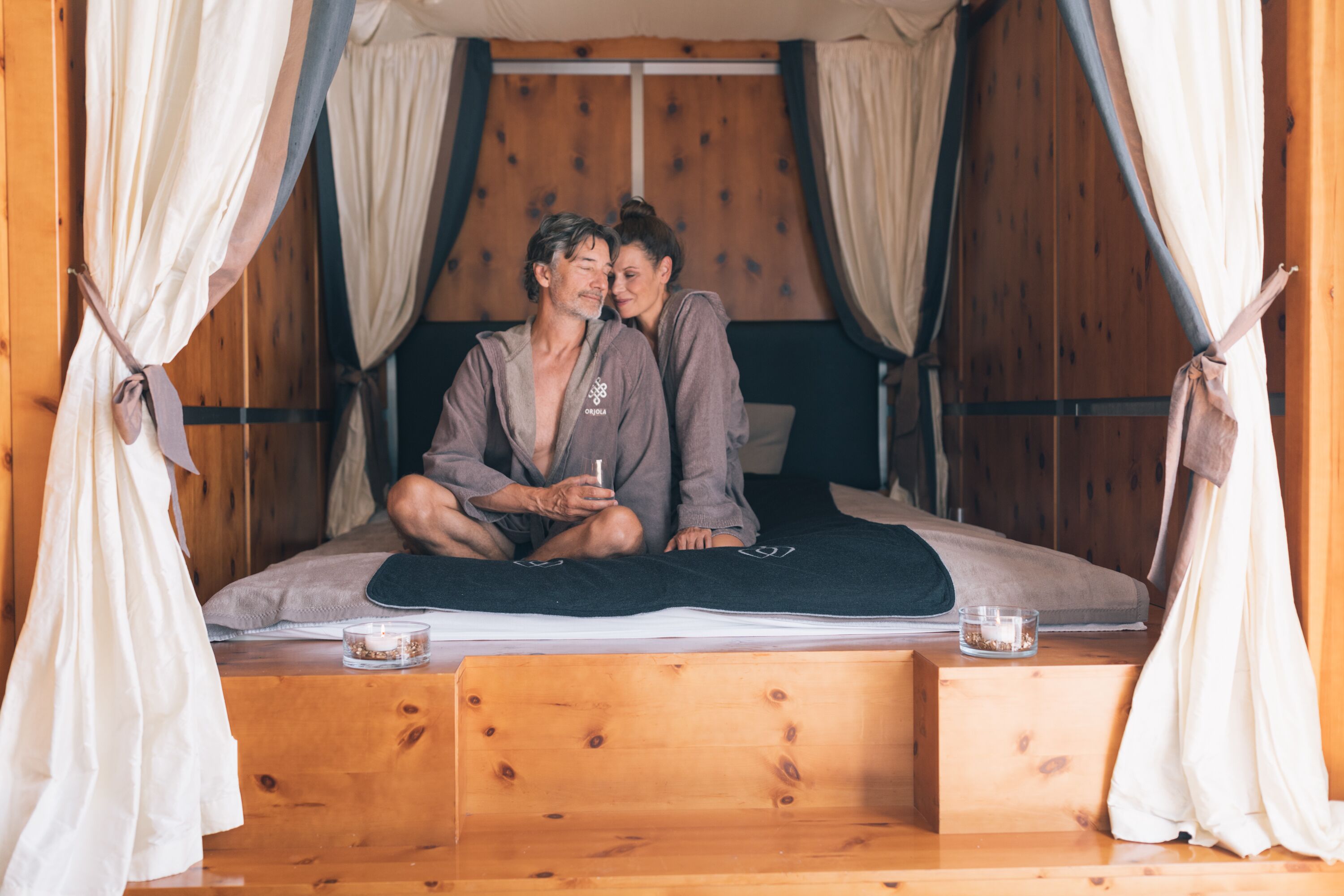 A wooden four-poster bed with a black bedspread, two pillows, and a glass candle on the side.