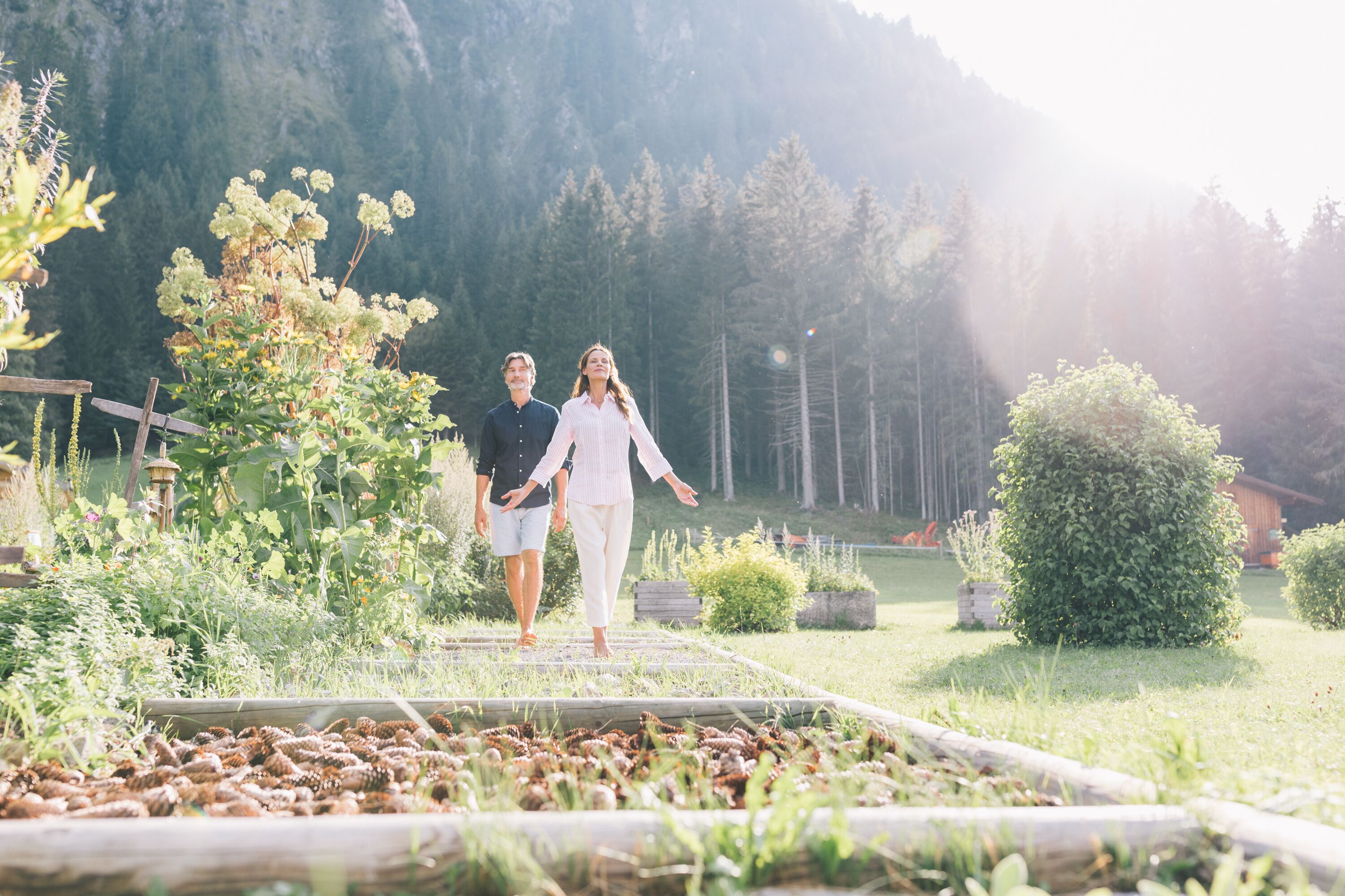 A couple walking on a wooden pathway surrounded by greenery and a mountainous backdrop.