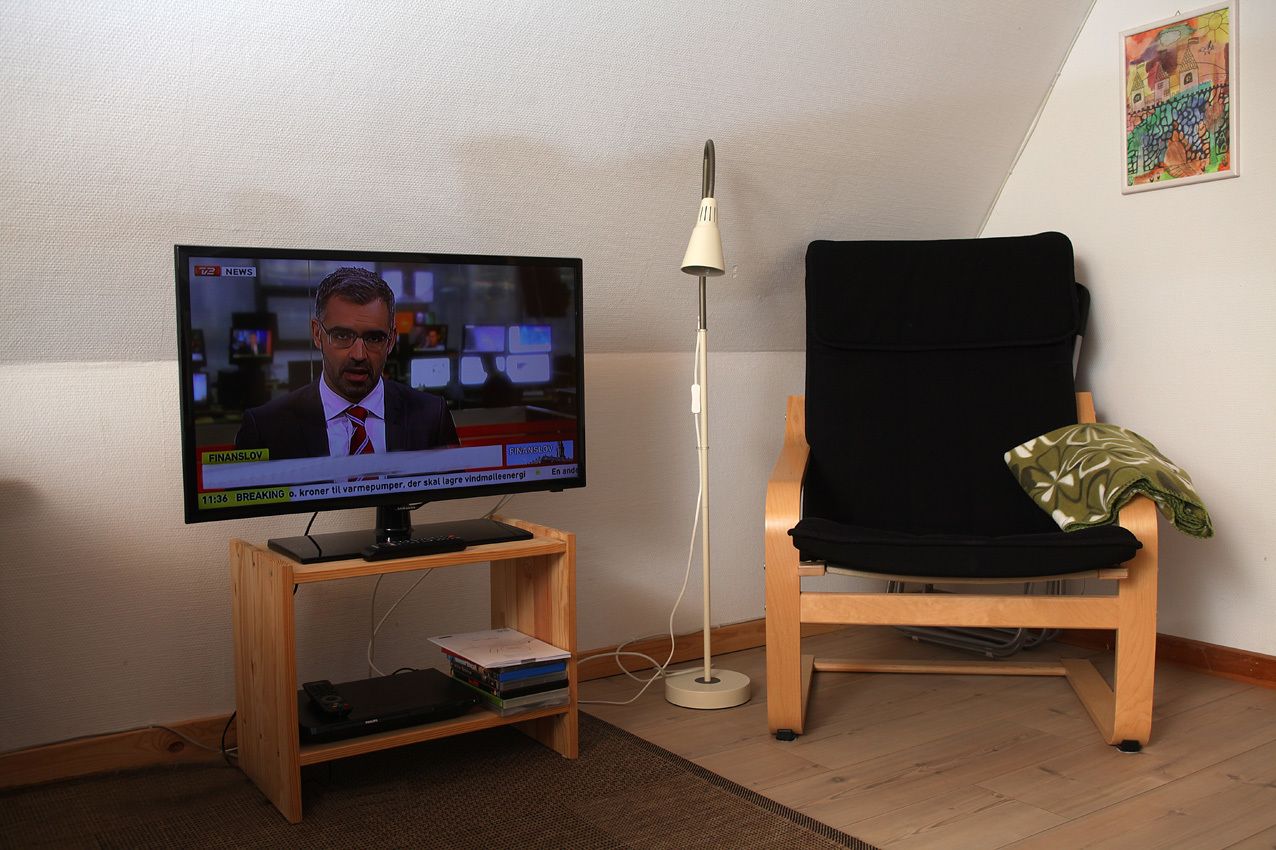 Un televisor sobre un soporte de madera, una silla negra y una lámpara de pie en una habitación con pared blanca y un cuadro colorido.