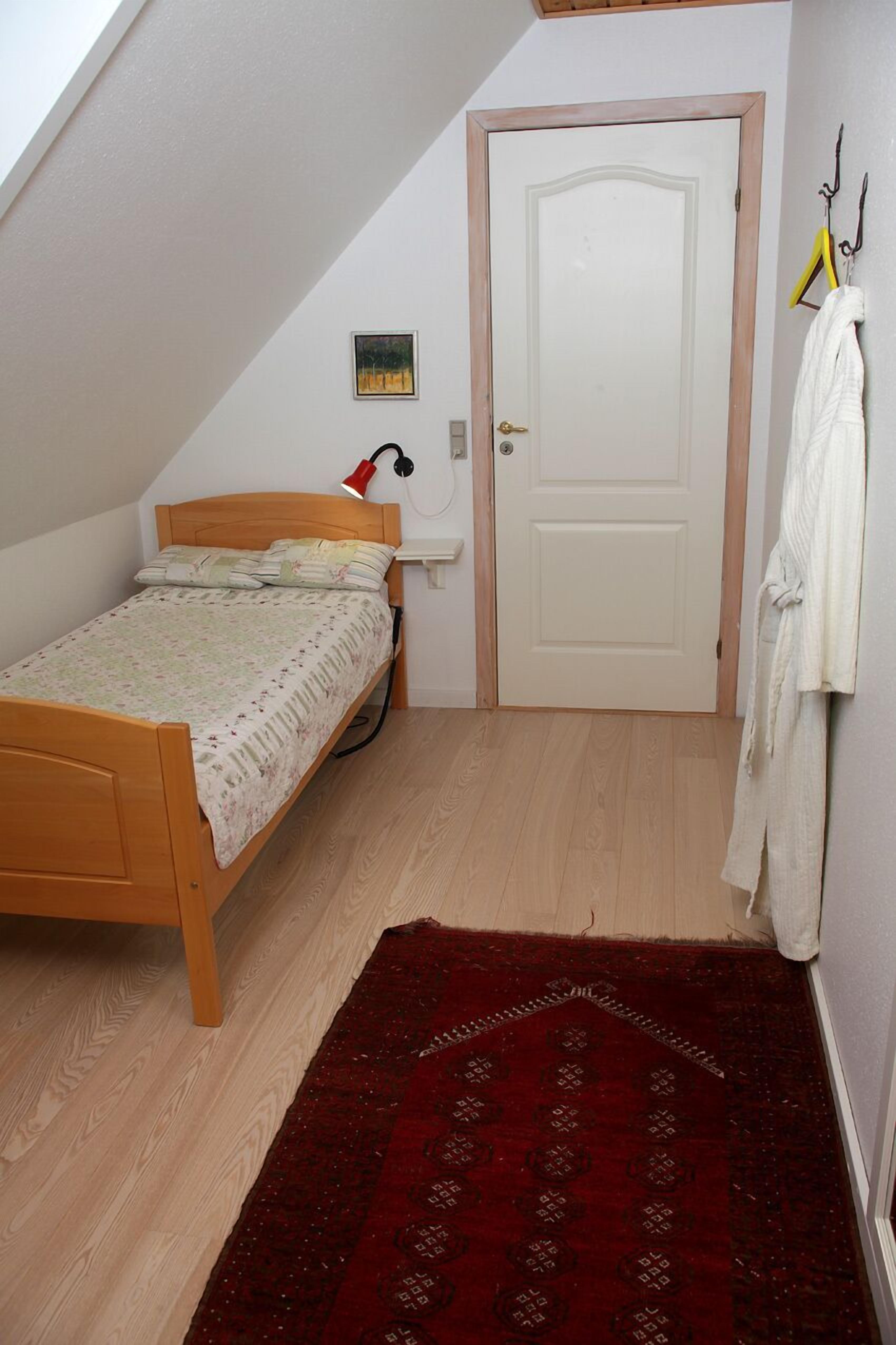 Un dormitorio pequeño y ordenado con una cama de madera, una alfombra roja, una puerta blanca y un cuadro pequeño en la pared.