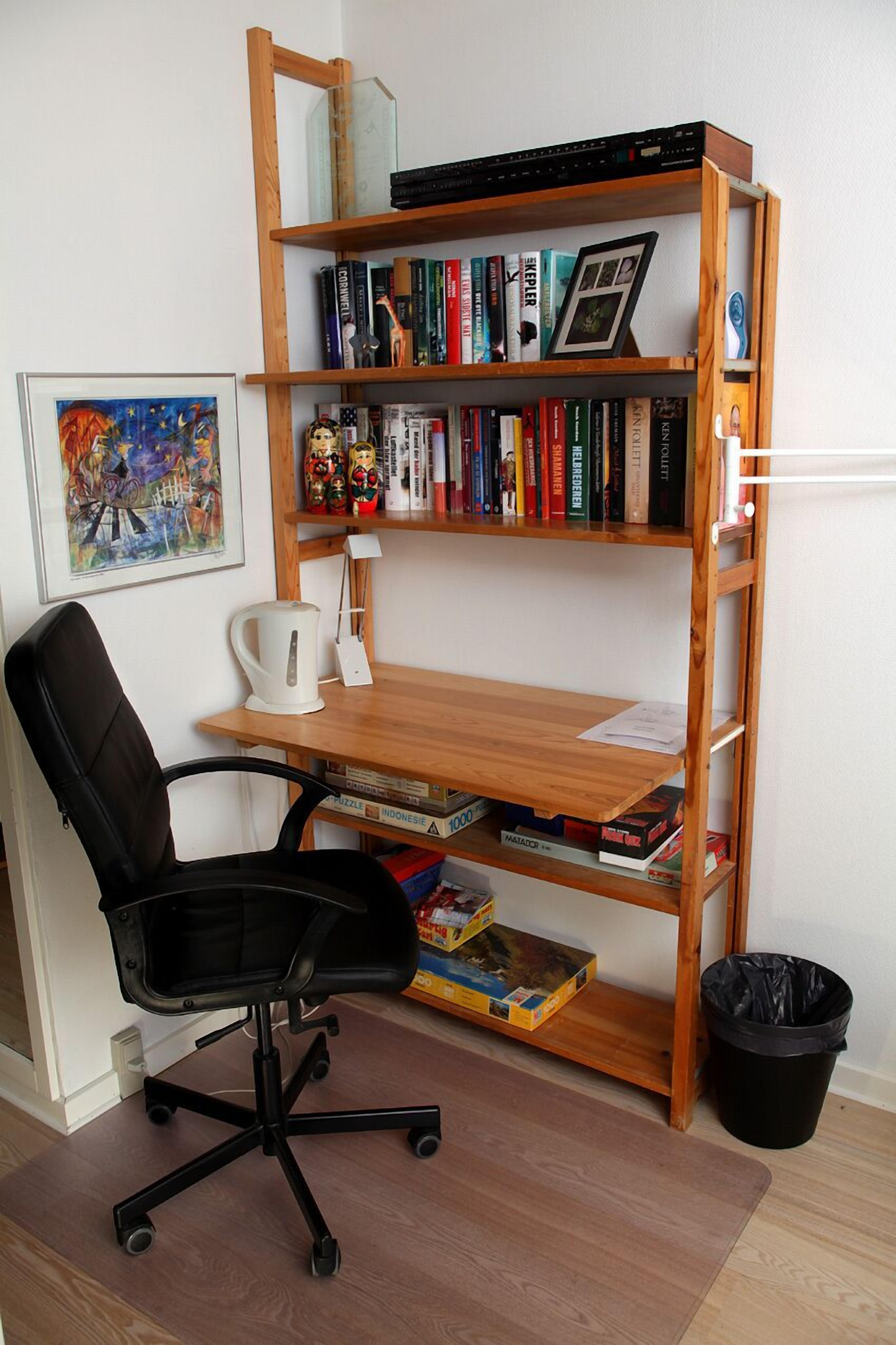Una habitación con un escritorio de madera, una silla de oficina negra, una estantería, un cuadro enmarcado y una cafetera.